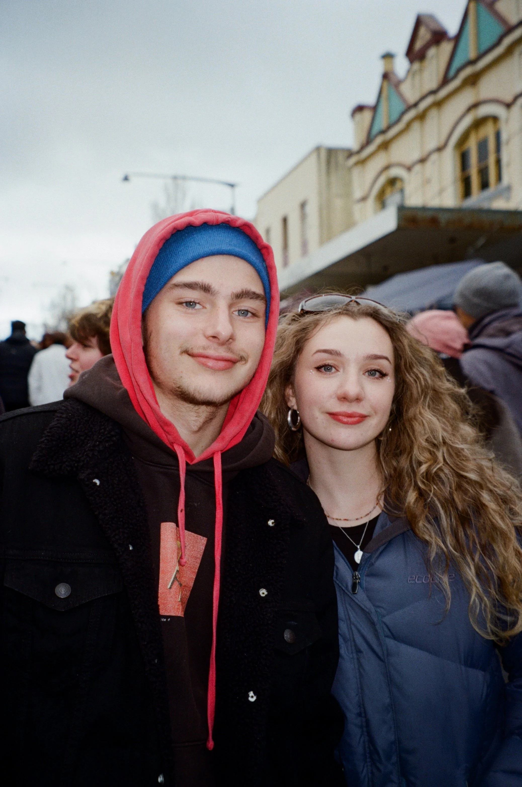 A young man with blue eyes, a slight smile, wearing a pink hoodie and blue beanie, and a young woman with curly hair, earrings, and a necklace, standing outdoors among a crowd of people in winter clothing near old buildings with decorative architectu