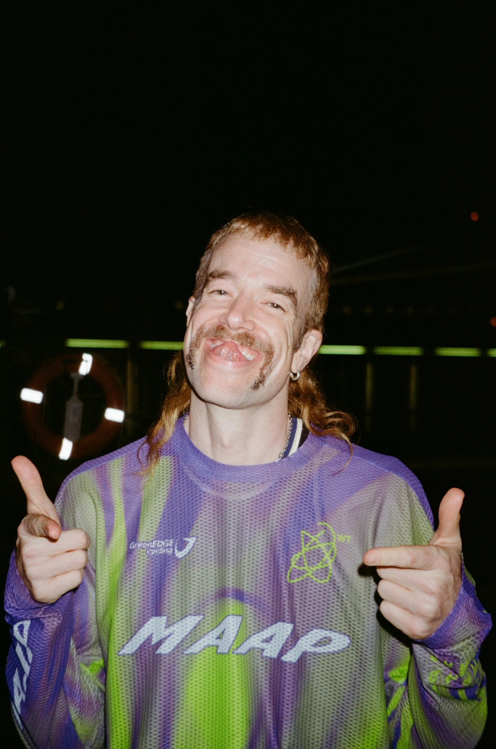 A person with a long, reddish-brown skullet, wearing a purple mesh sports jersey with bright green accents, is smiling and giving a finger guns motion with both hands. The background appears to be outdoor at a pub/bar