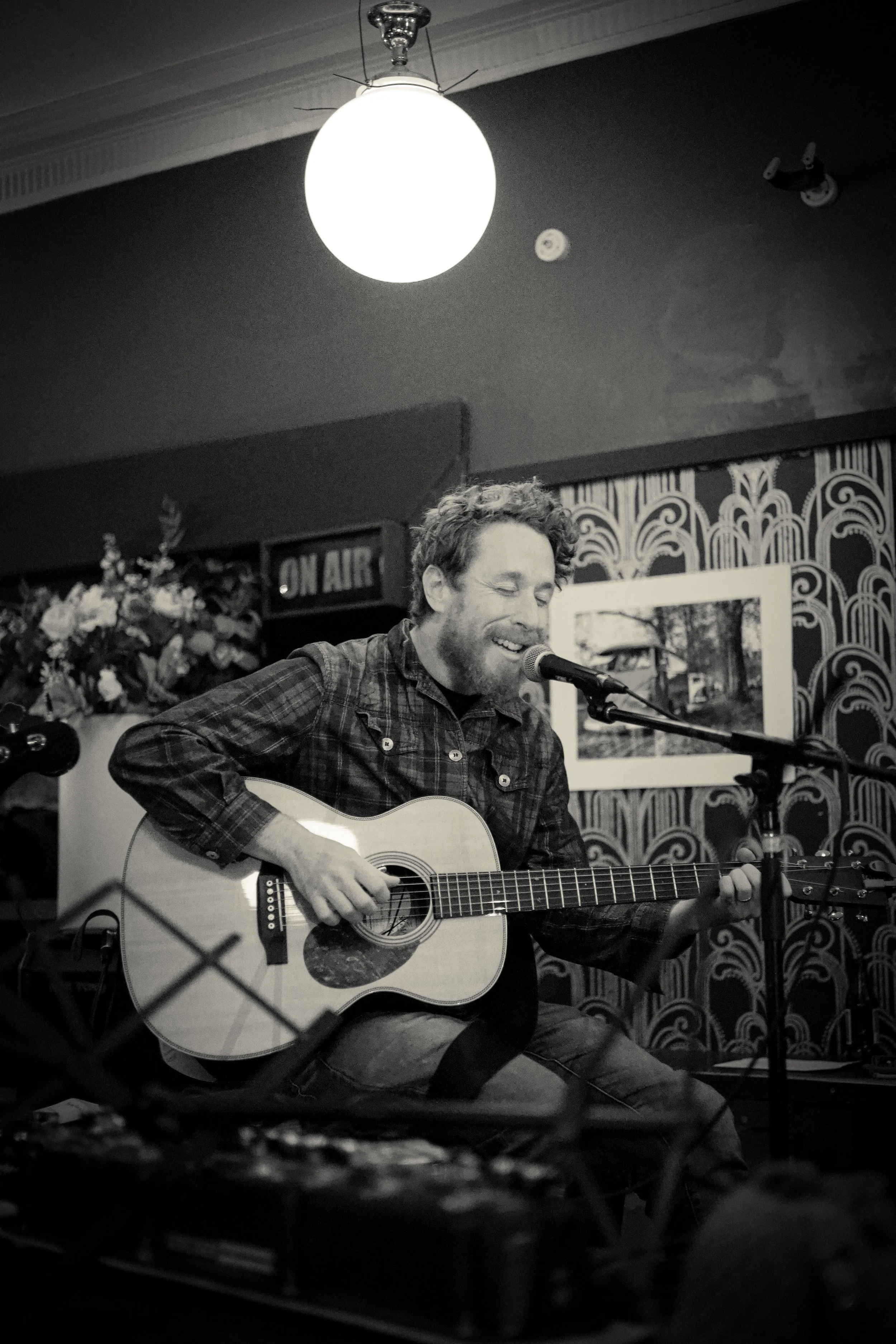 A man with curly hair and glasses playing an acoustic guitar and singing into a microphone in a cozy indoor setting with patterned wallpaper and flowers on a table behind him.