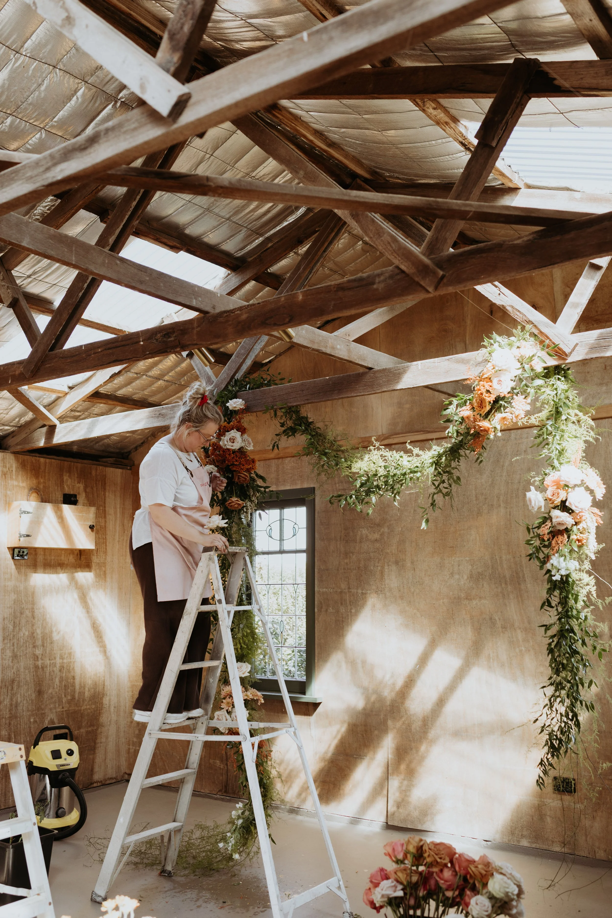 A woman standing on a ladder decorating a room with flowers and greenery for an event, with wooden beams and a window in the background. Krissy from Studio Zgela on site installing a floral installation at Chapter Barn, South Gippsland.