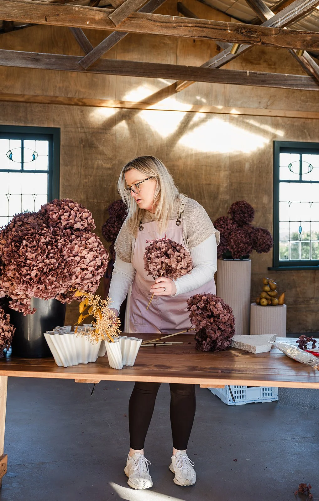 A woman arranging pink hydrangea flowers in a black vase inside a rustic room. There are large windows, wooden ceiling beams, and a wooden table with flower arrangements and supplies at Chapter Barn.