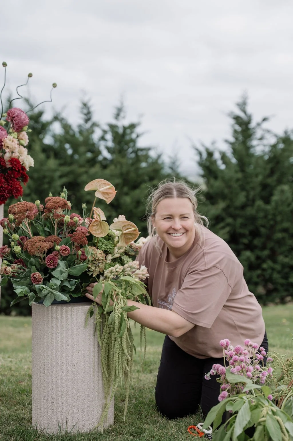 A woman with blonde hair is kneeling on the grass next to a large flower arrangement in a tall beige vase, smiling at the camera. The arrangement includes pink, red, and cream-colored flowers, with green foliage. In the background, there are trees and a cloudy sky.