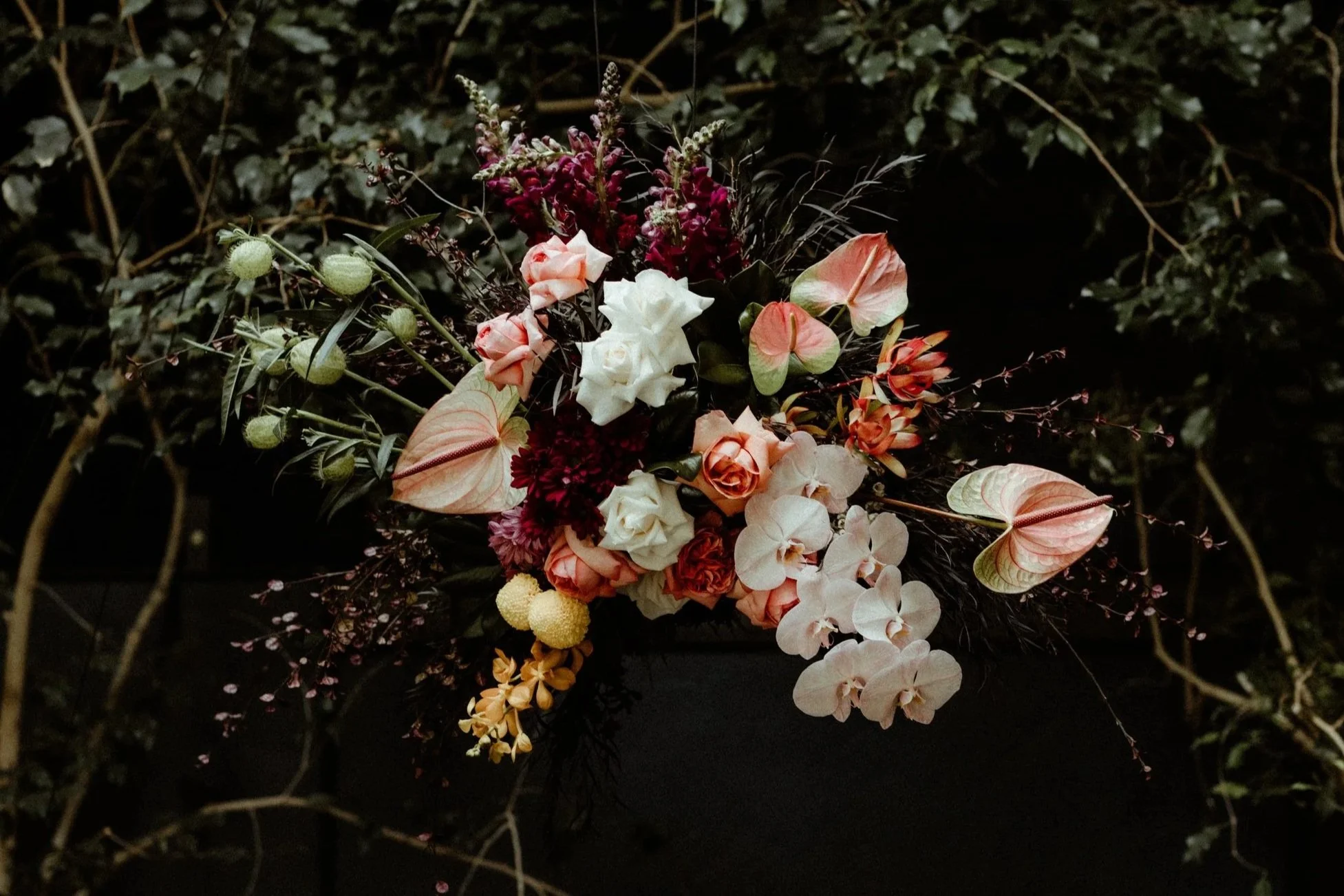 A floral arrangement with a variety of colorful flowers including roses, anthuriums, and other blooms, set against a dark background surrounded by green foliage.