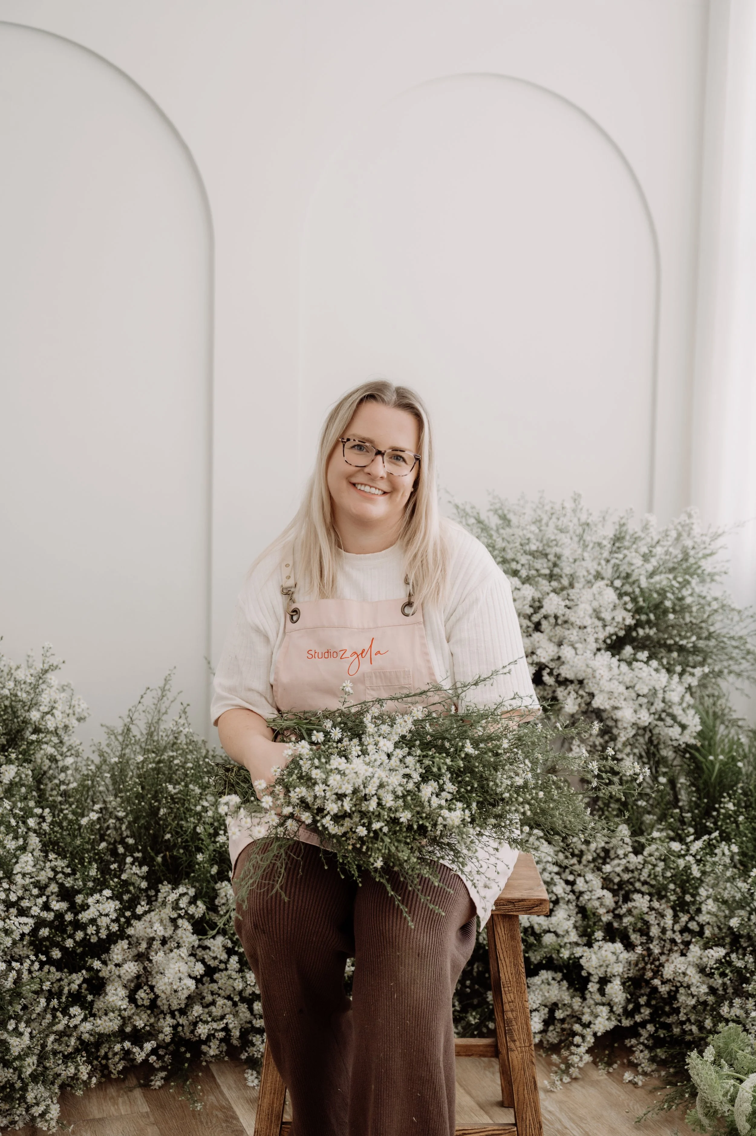 A woman with glasses and blonde hair sitting on a wooden stool in front of a white wall with floral arrangements of white flowers and greenery surrounding her.