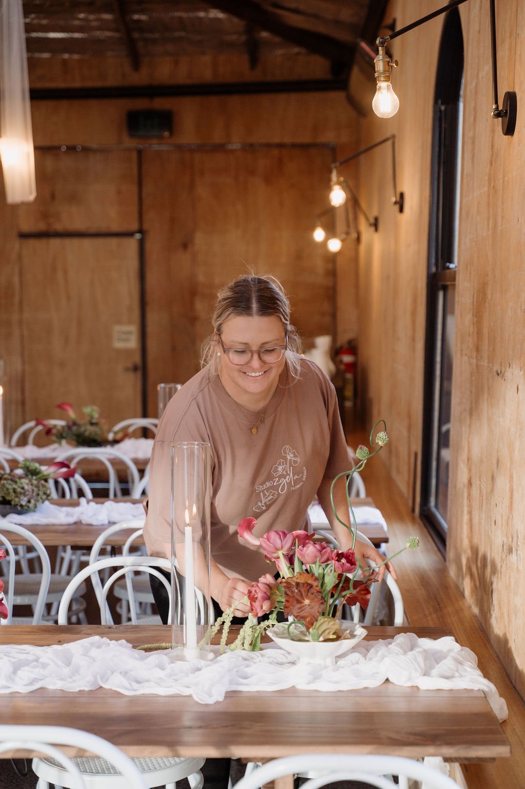 A woman arranging pink and green floral centerpiece on a wooden table in a rustic wooden room with warm lighting.