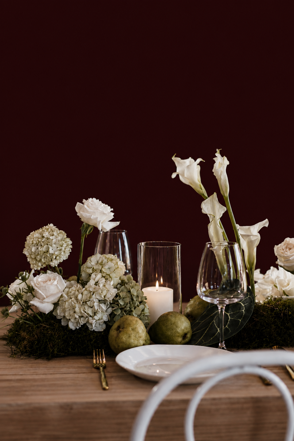 Elegant table setting with white flowers, a lit candle, pears, glassware, and a white plate on a wooden table against a dark background.