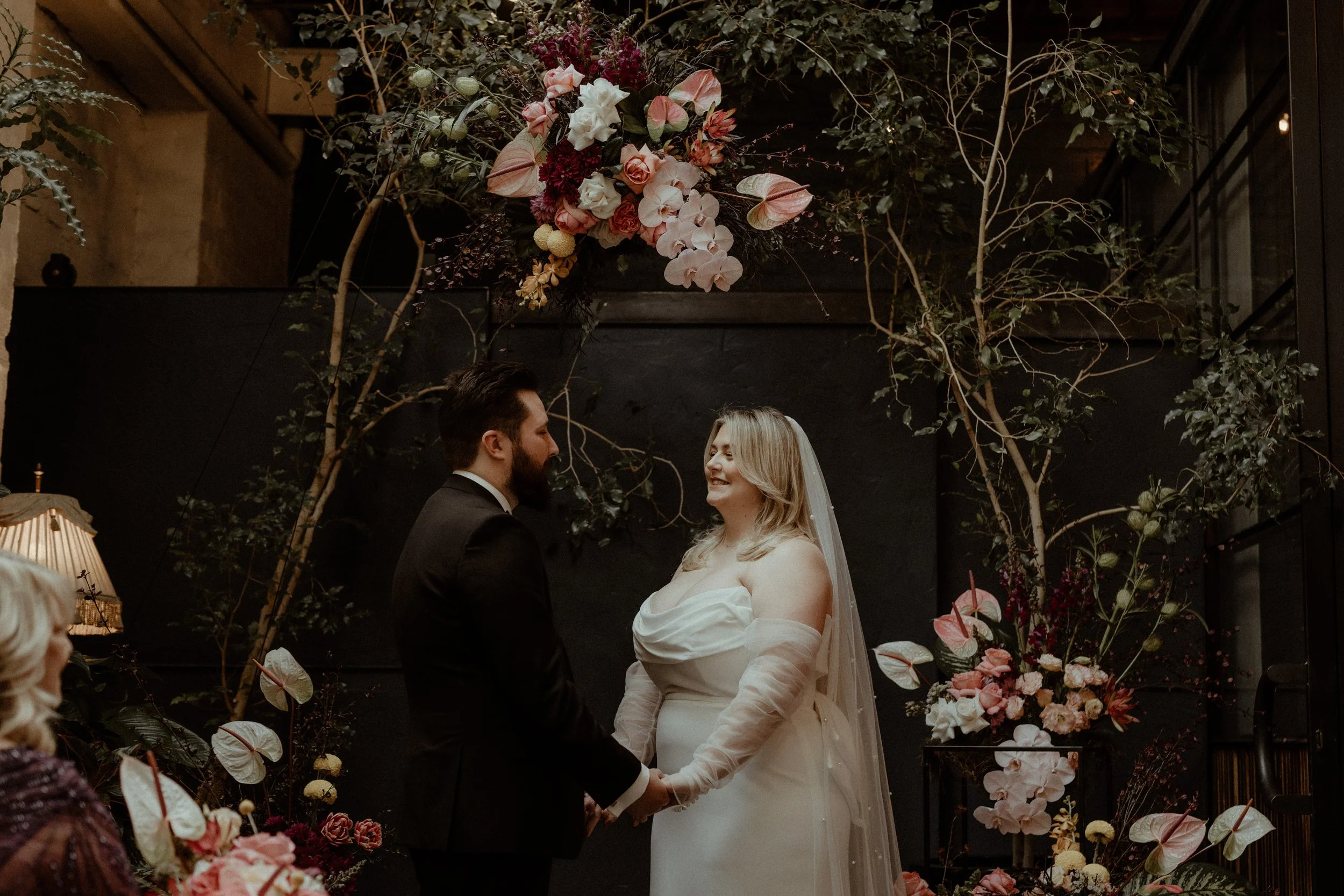A bride and groom holding hands during their wedding ceremony, standing in front of a floral backdrop with pink and white flowers and greenery.