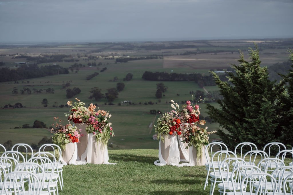 Outdoor wedding ceremony setup with white chairs and colorful flower arrangements on white vases, overlooking green fields and distant farmland at chapter Barn.