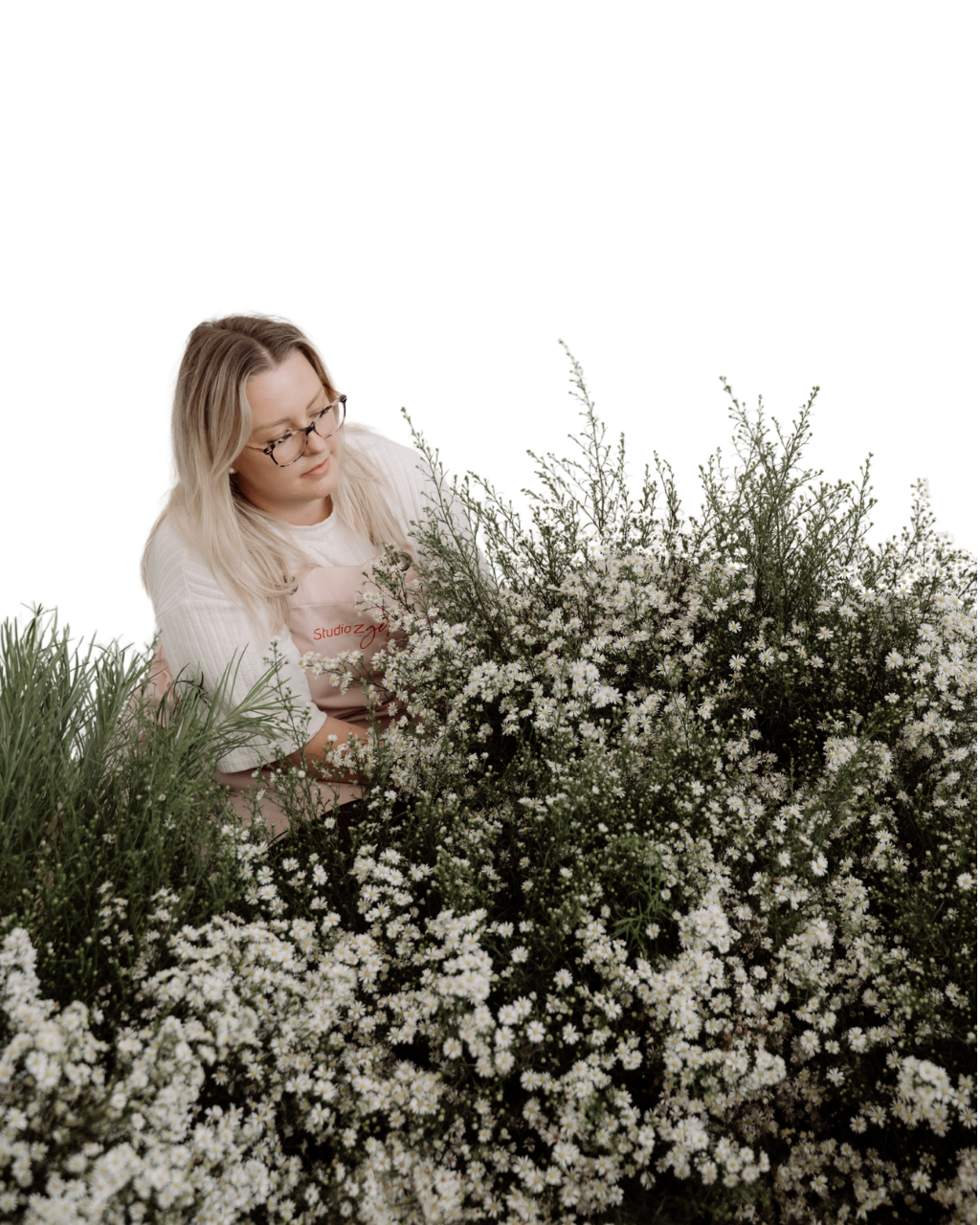 A woman with blonde hair and glasses crouches among white daisies and greenery, looking at the flowers.