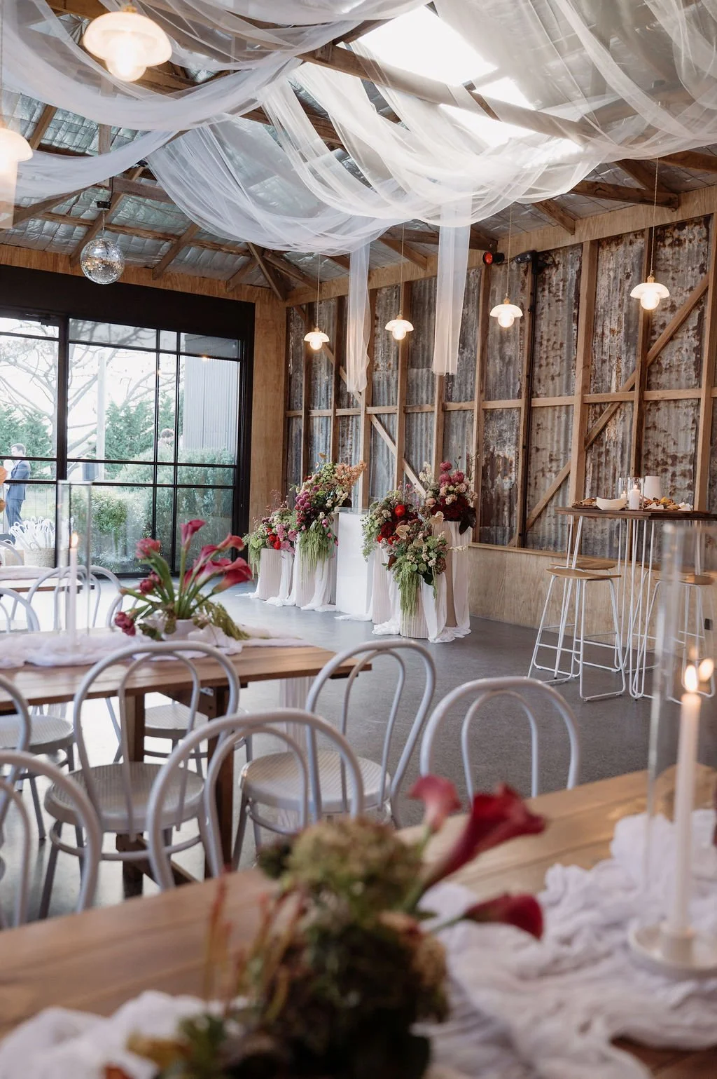 Interior of a rustic wedding reception venue with hanging fabric and lights, floral centerpieces, and wooden furniture.