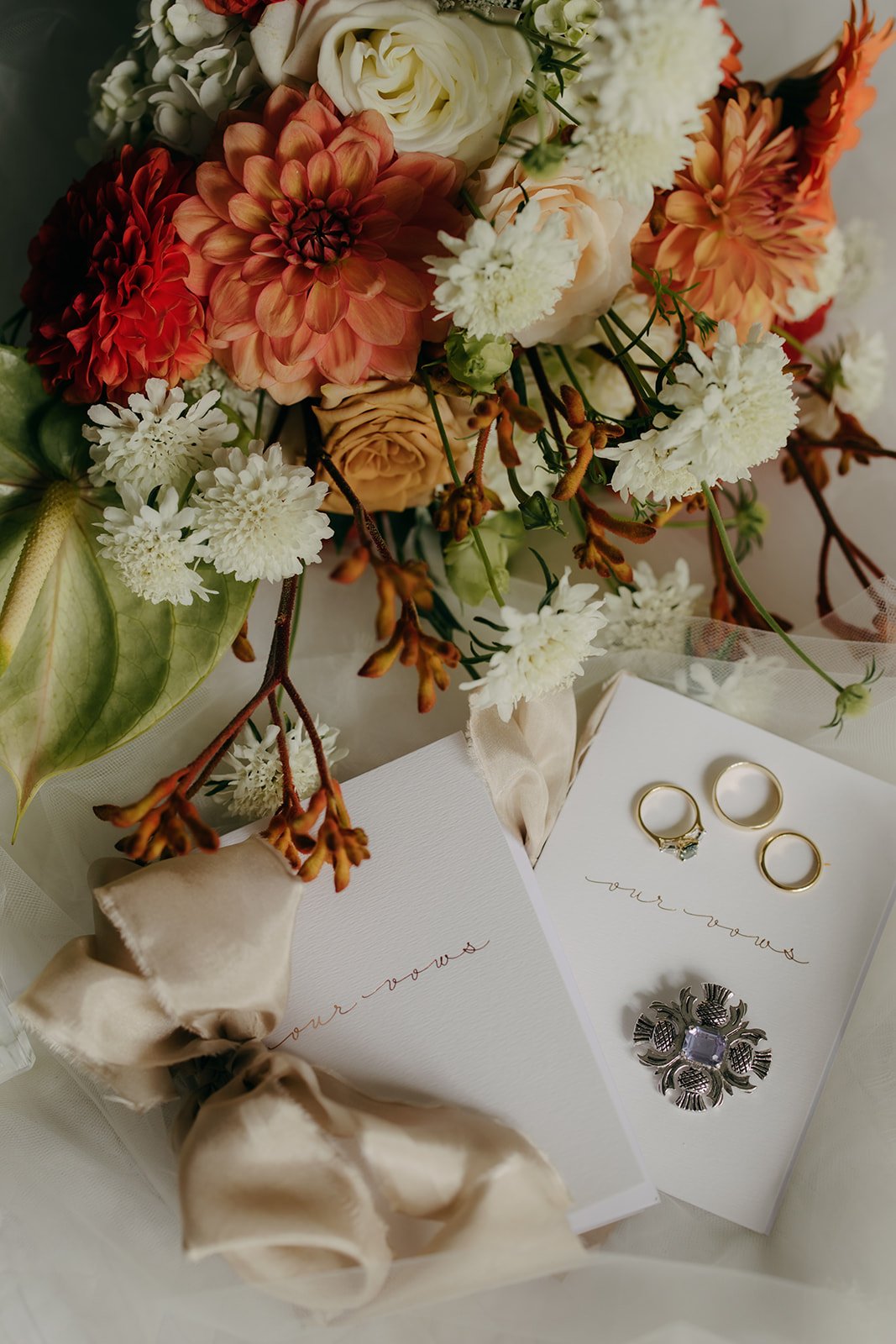A bouquet of white, orange, and peach flowers including dahlias, roses, and chrysanthemum, with green leaves, accompanying wedding rings and jewelry, laid on a white surface with notes that say 'our vows'.