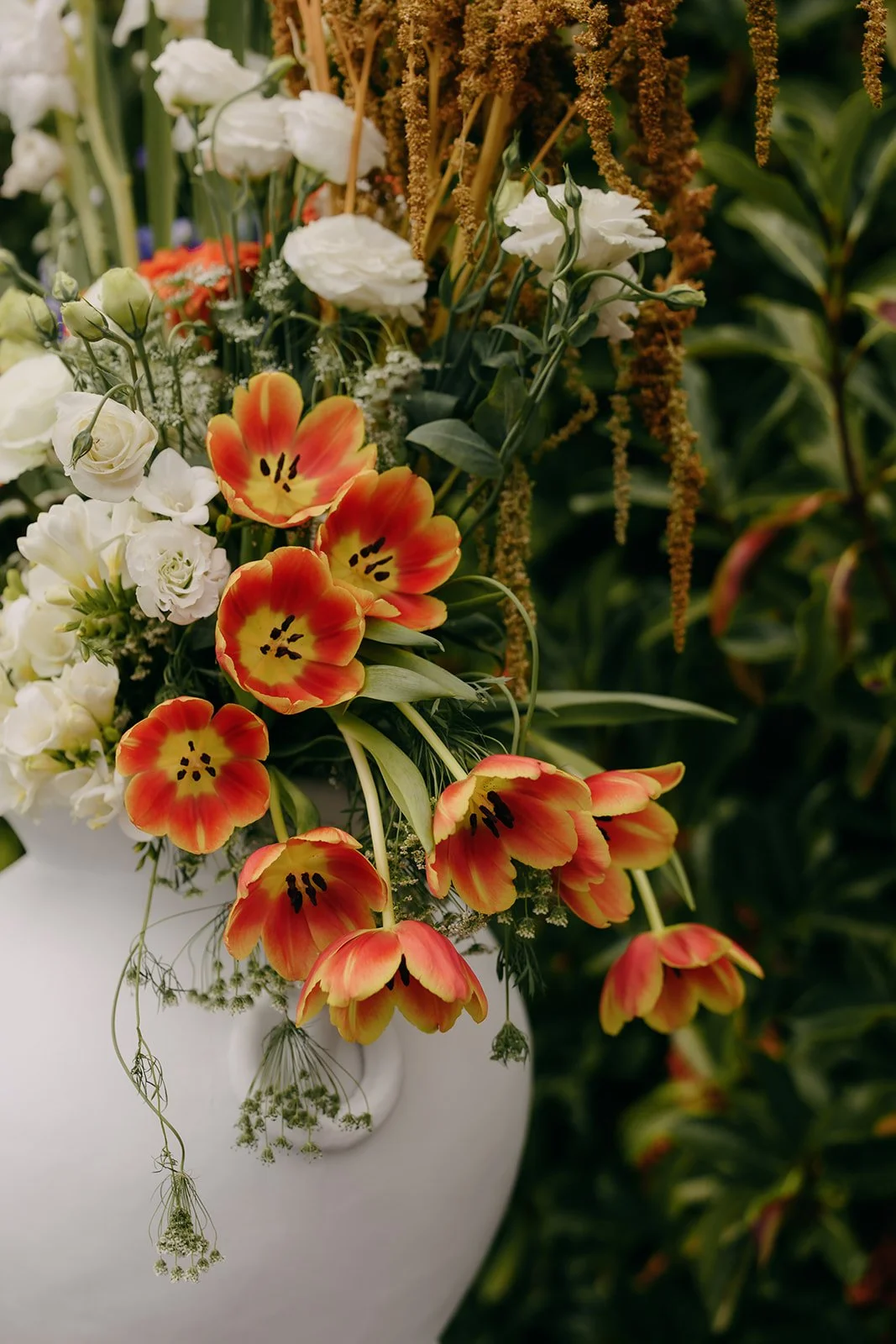 A white vase holding a colorful arrangement of flowers including orange and yellow tulips, white lisianthus, and other small white and purple flowers with greenery, set against a blurred green leafy background.