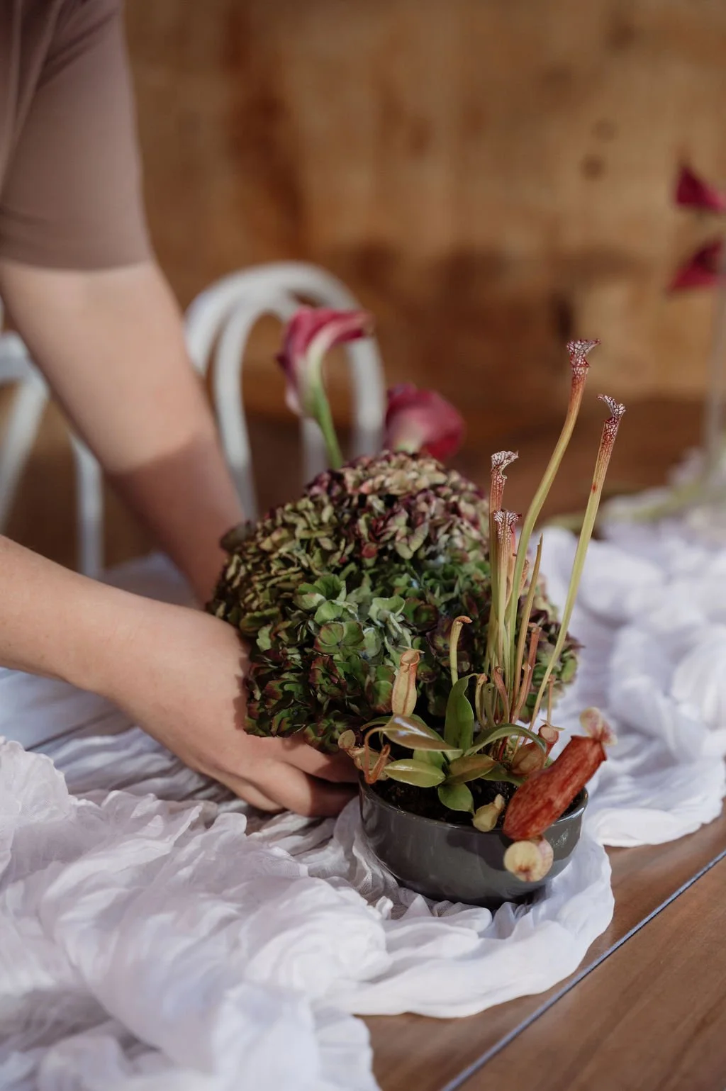 Person arranging a floral centerpiece with hydrangeas, roses, and pitcher plants on a white fabric tablecloth.