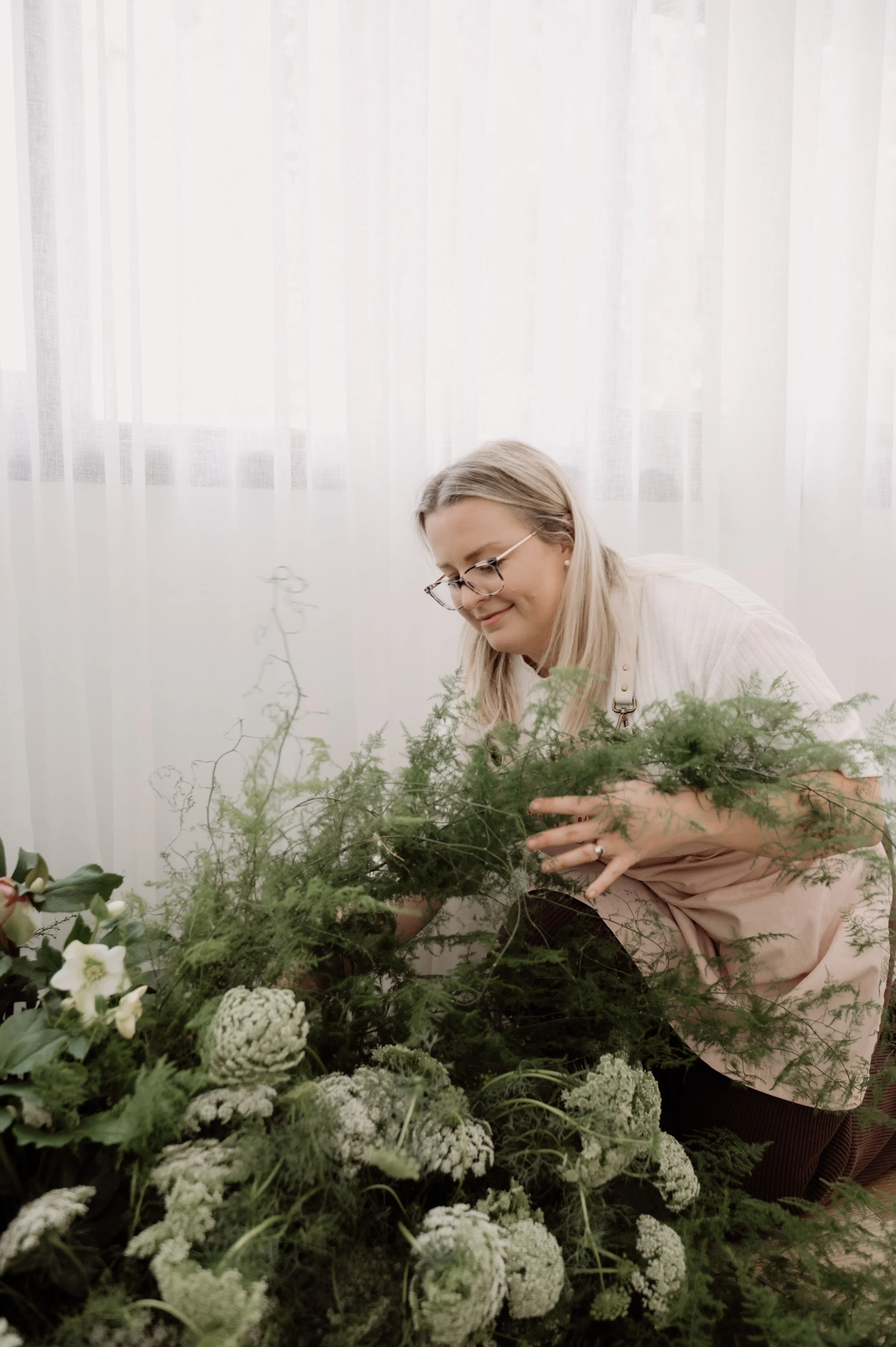 Woman with blonde hair, glasses, wearing a light-colored top, arranging white flowers and greenery indoors with a sheer white curtain background.