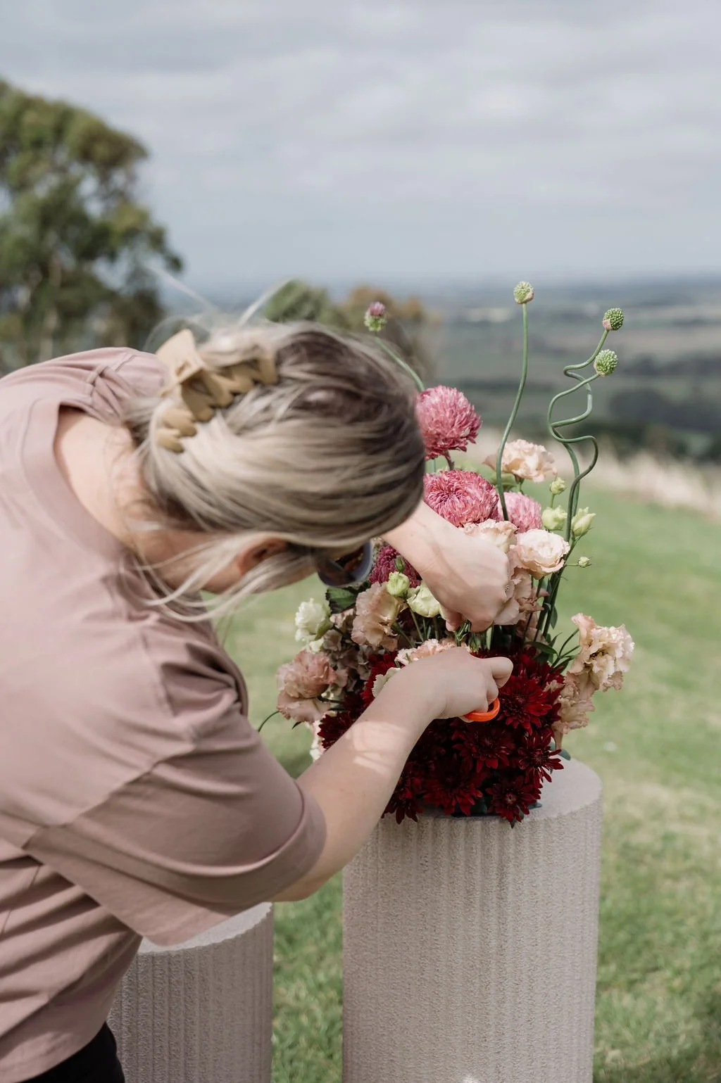 A woman with blonde braided hair arranging a bouquet of pink and red flowers outdoors on a white pedestal, with a scenic landscape and cloudy sky in the background.