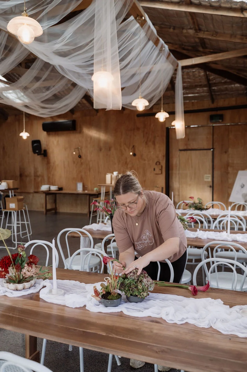 A woman arranging flowers on a decorated table in a rustic indoor setting with wooden walls and ceiling, white chairs, and soft lighting hanging from the ceiling.