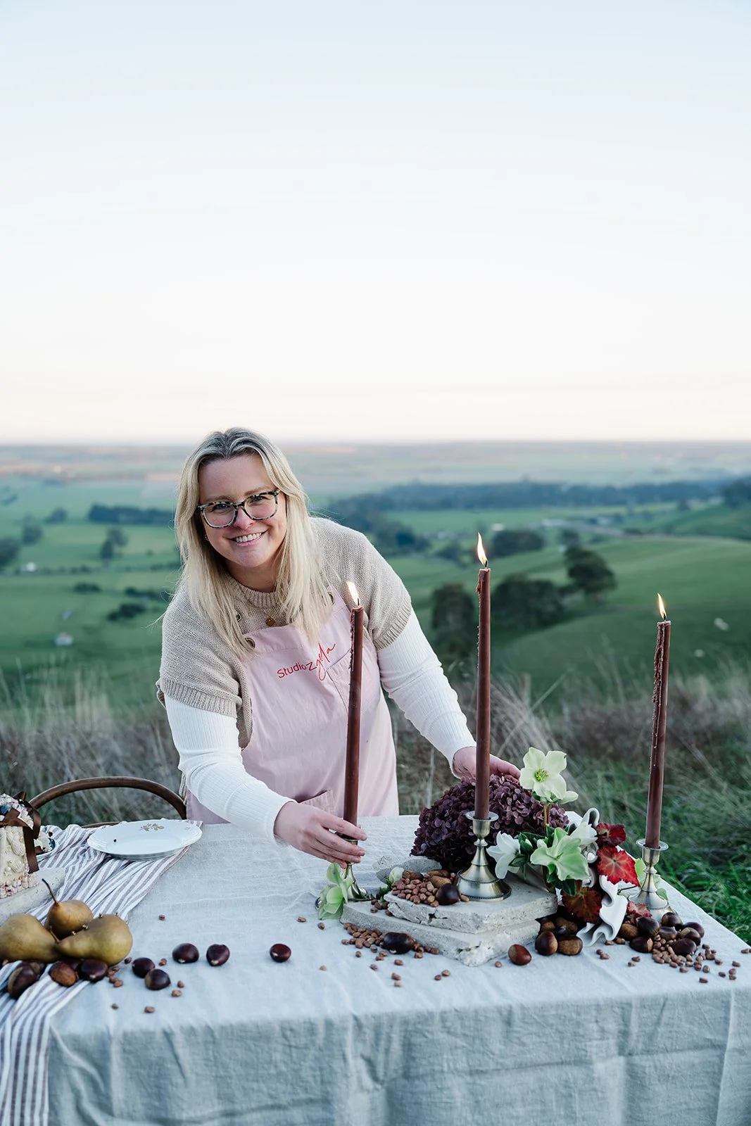 A woman with blonde hair and glasses smiling at a table set outdoors with candles, flowers, and chocolates, overlooking a grassy landscape with fields and trees in the distance at Chapter Barn.