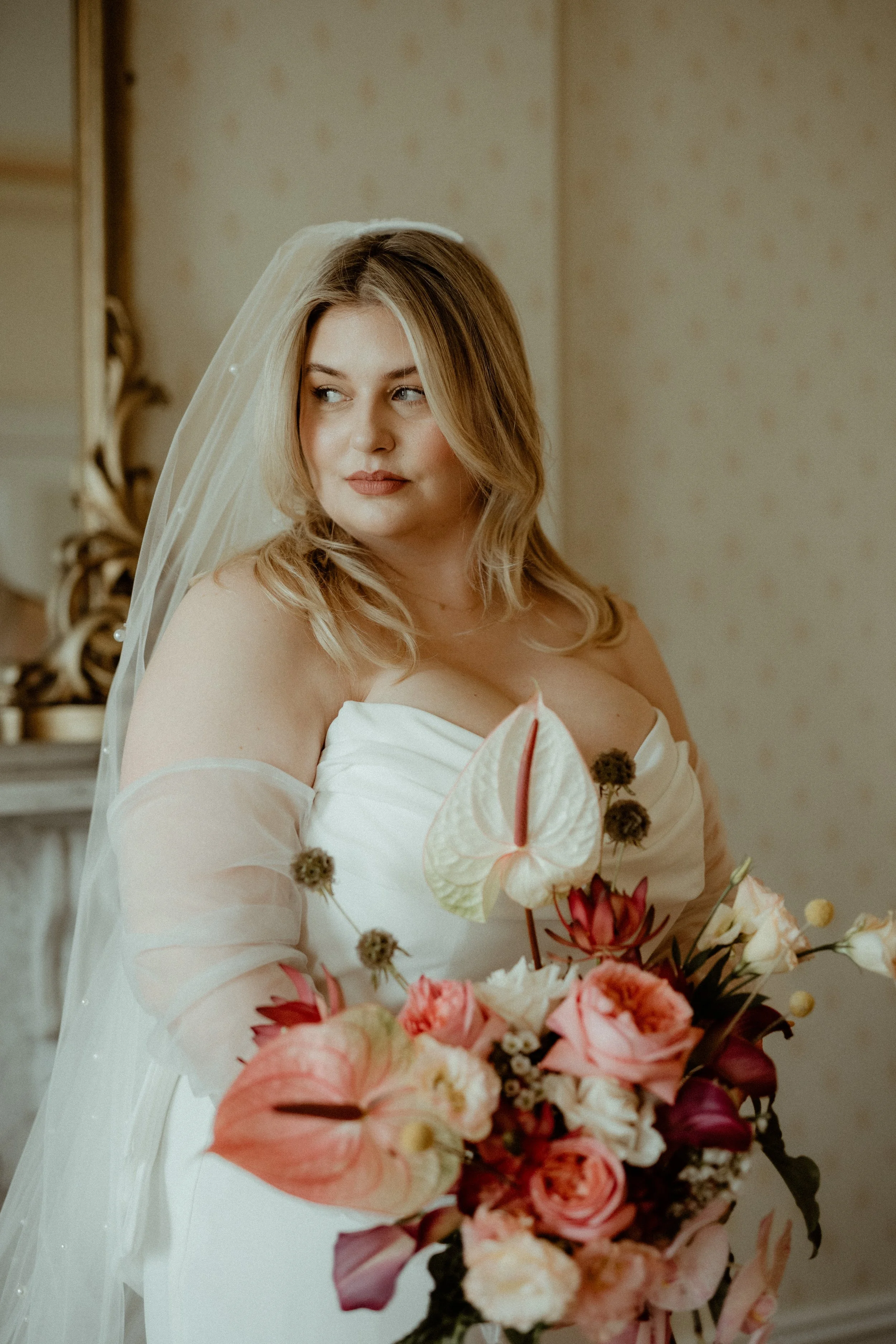 A bride with blonde hair in a wedding dress holding a colorful bouquet of flowers, standing indoors.