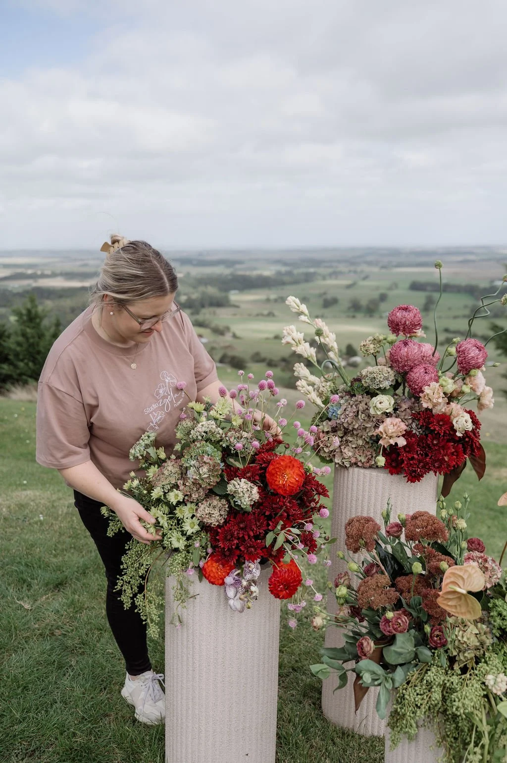 A woman arranging a colorful flower bouquet outdoors on a grassy hill with a scenic landscape and cloudy sky in the background.
