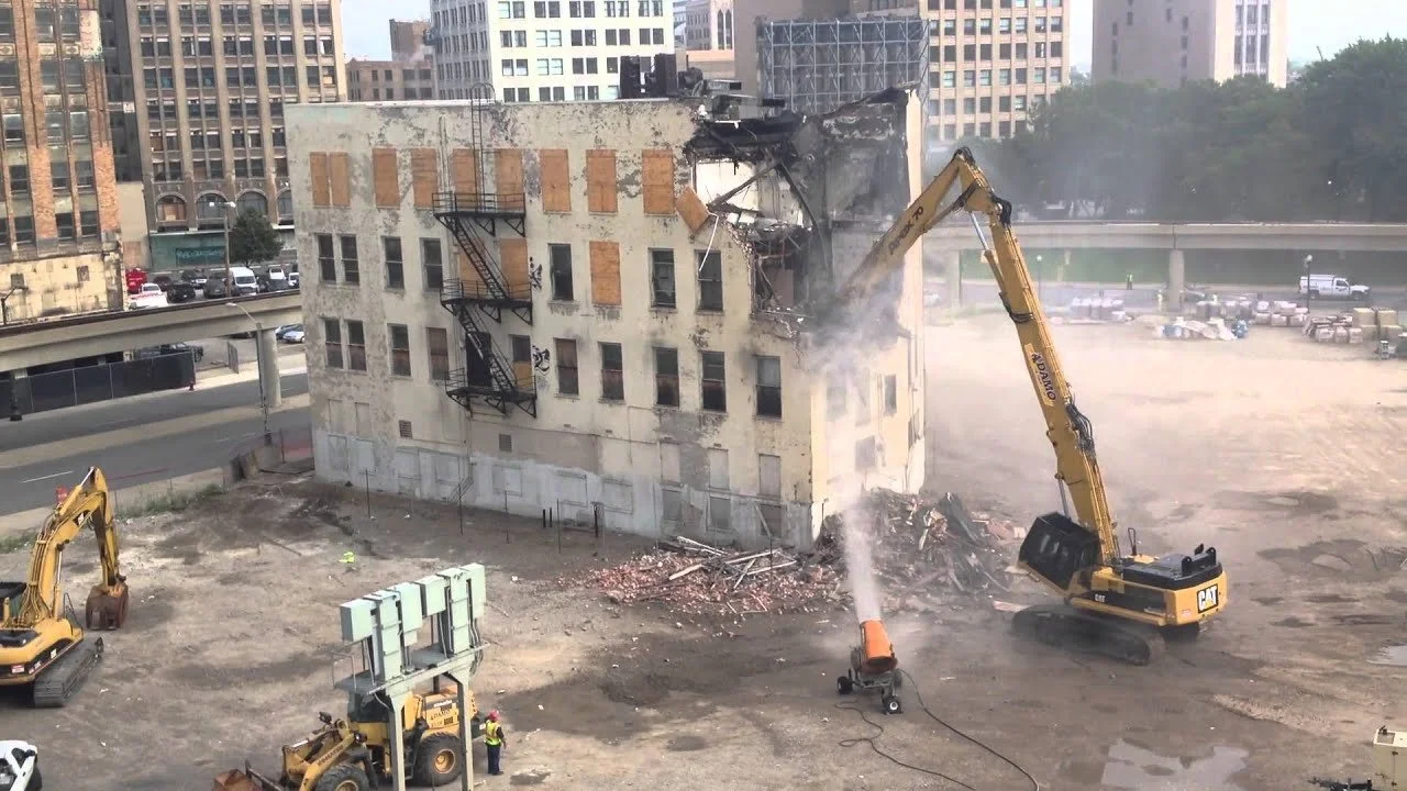Demolizione di un edificio di cinque piani con escavatori gialli. Una parete viene abbattuta, creando polvere e detriti.