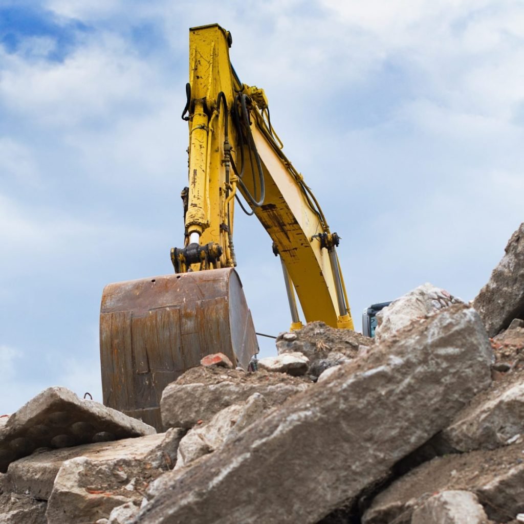 Cantiere con escavatore giallo che lavora su una pila di rocce e detriti sotto un cielo con nuvole.