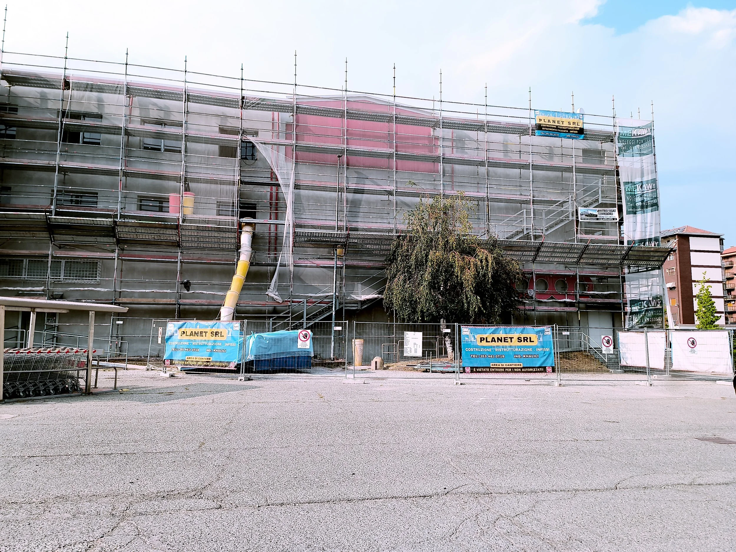 Edificio in costruzione circondato da impalcature e pannelli di rete, con cartelli pubblicitari che indicano la ditta esecutrice e restrizioni di accesso, sotto un cielo parzialmente nuvoloso.