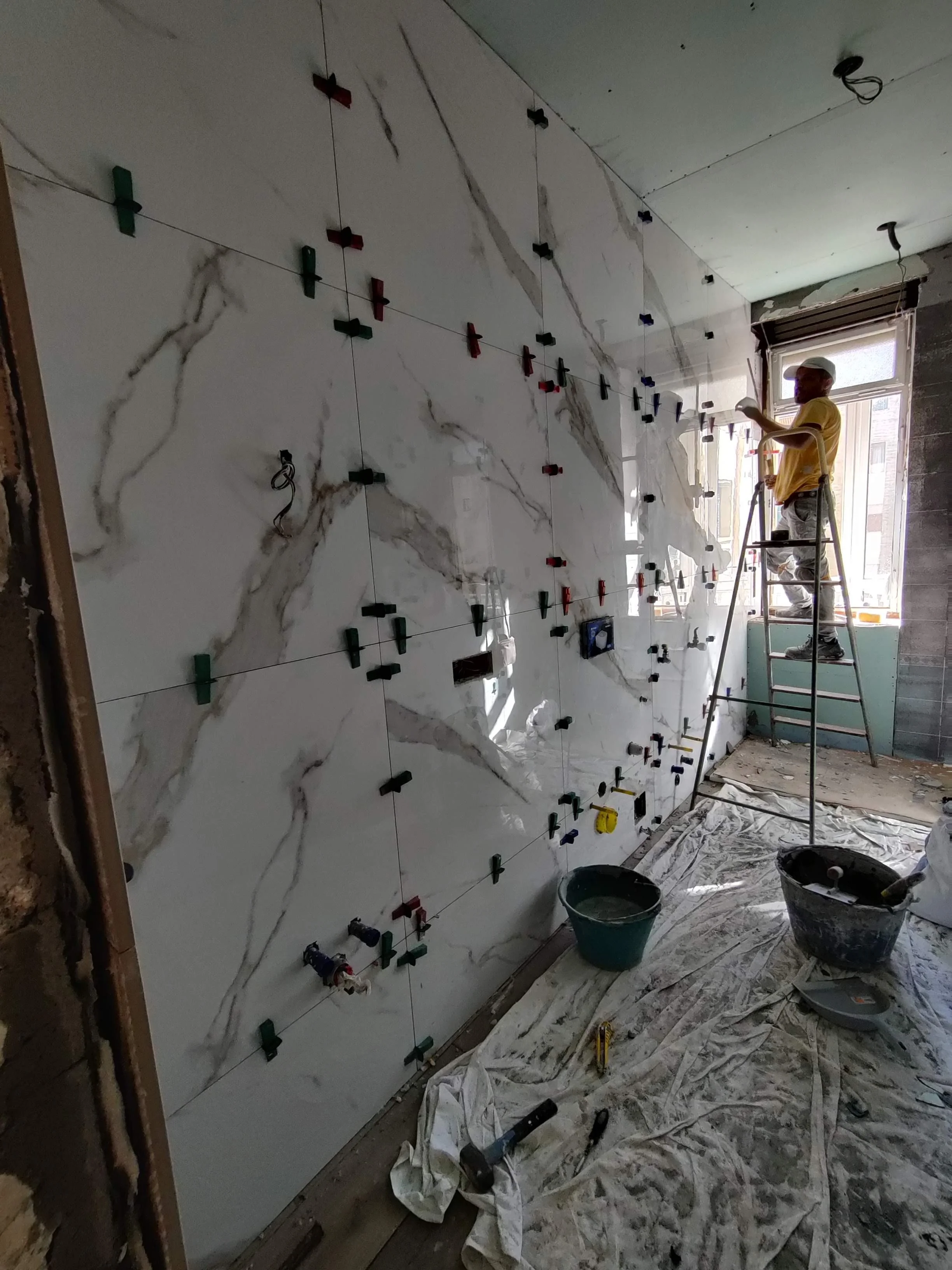 Una persona su una scala sta installando piastrelle di marmo bianche con venature grigie su una parete interna di una stanza in ristrutturazione. Sul pavimento ci sono teli e attrezzi da costruzione.