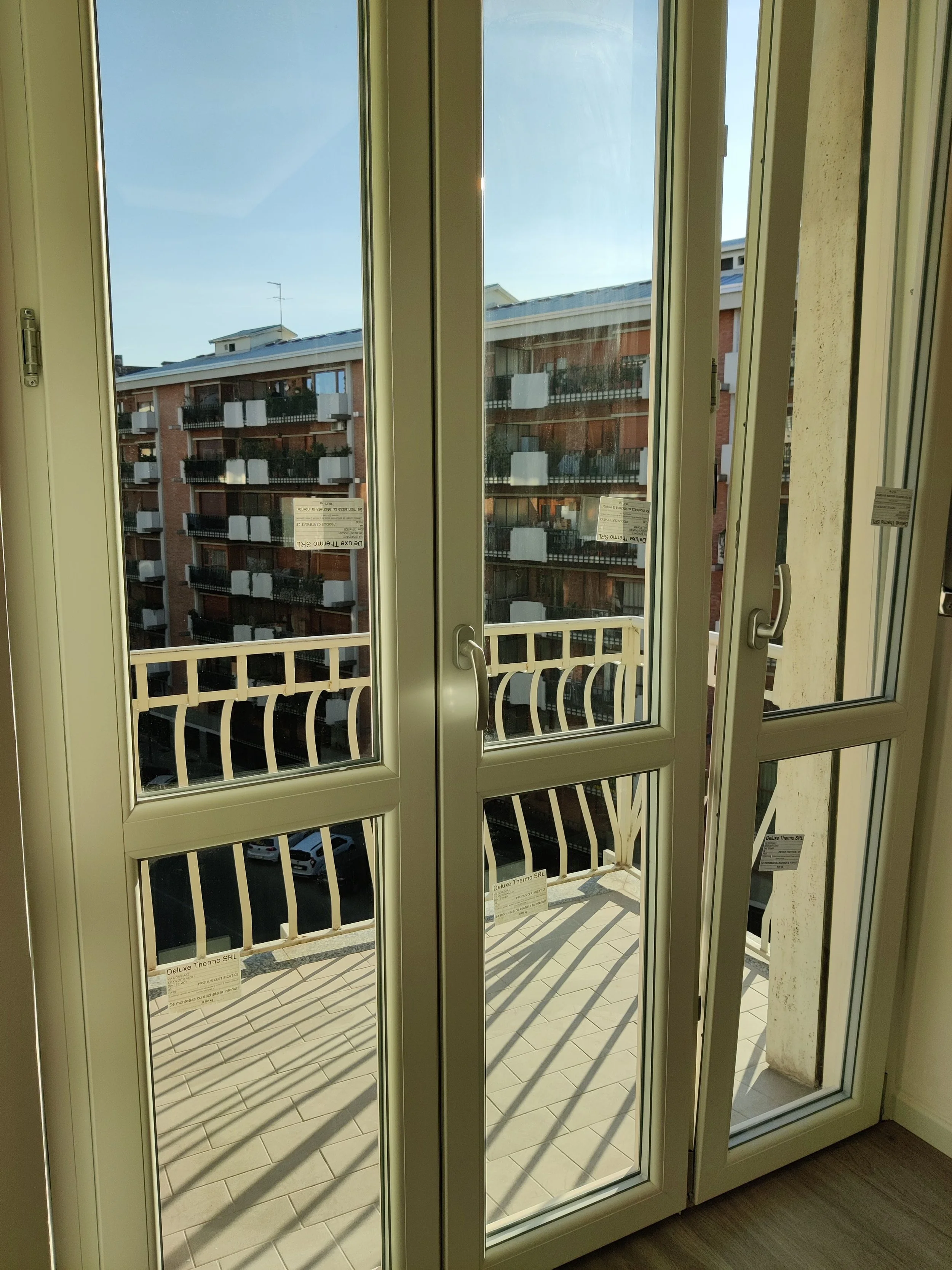 Immagine di una finestra con ante di vetro che si apre su un balcone con ringhiera bianca. Sul balcone ci sono dei cartelli con scritte e un'ombra di ringhiera proiettata sul pavimento.