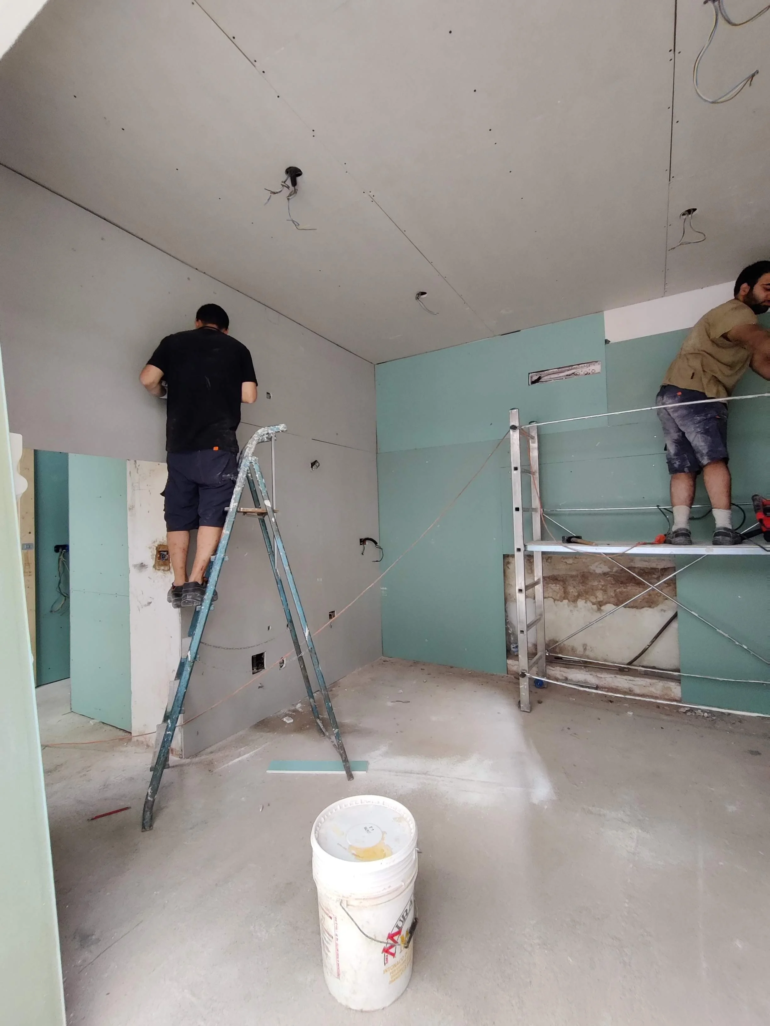 Due persone stanno installando le pareti e il controsoffitto in una stanza in fase di ristrutturazione, con strumenti e materiali da lavoro.