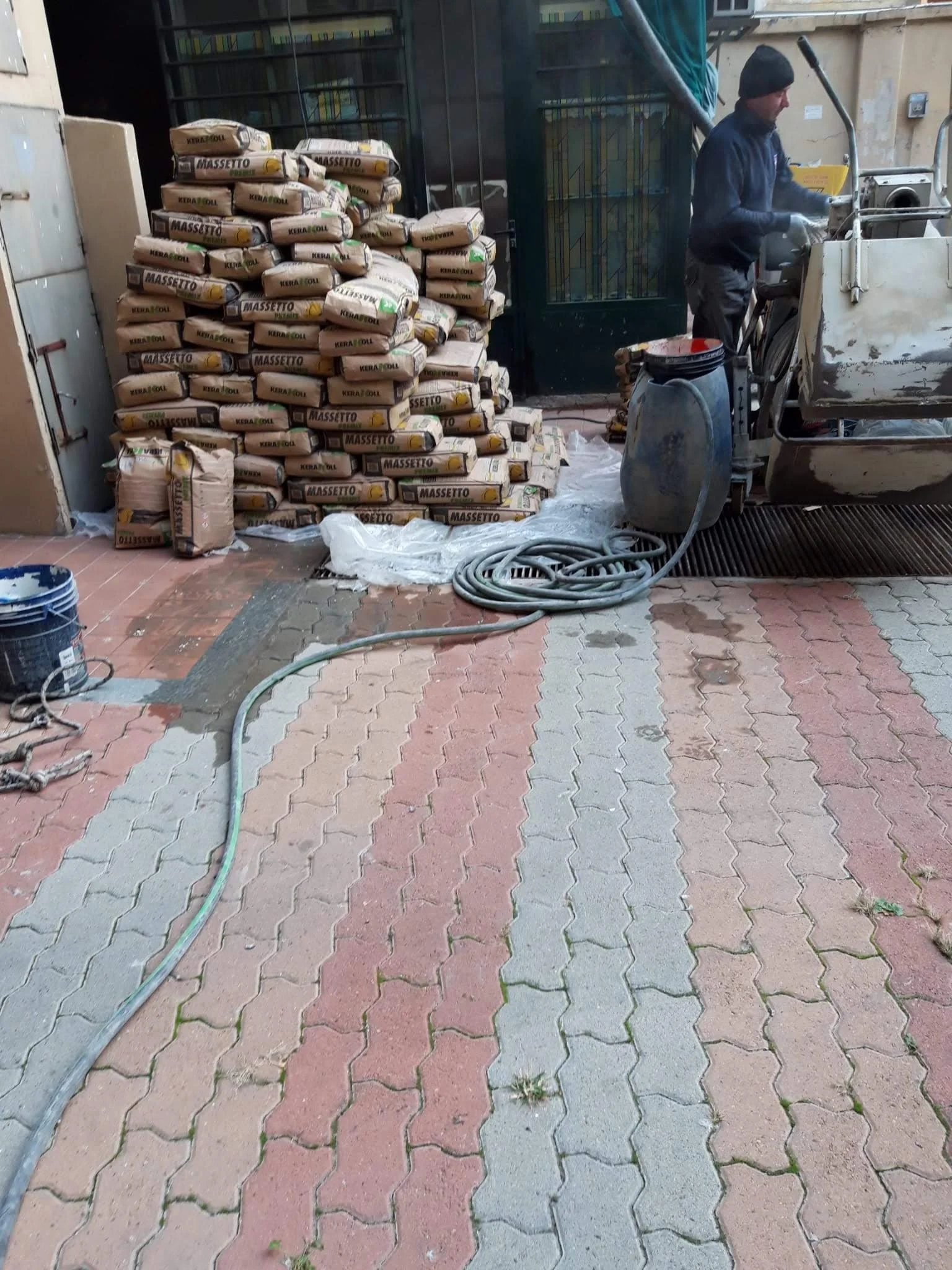 Un operatore sta utilizzando una macchina per il taglio o la lavorazione di materiali, con sacchi di cemento o simili accatastati sullo sfondo, situato in un'area esterna e asfaltata.