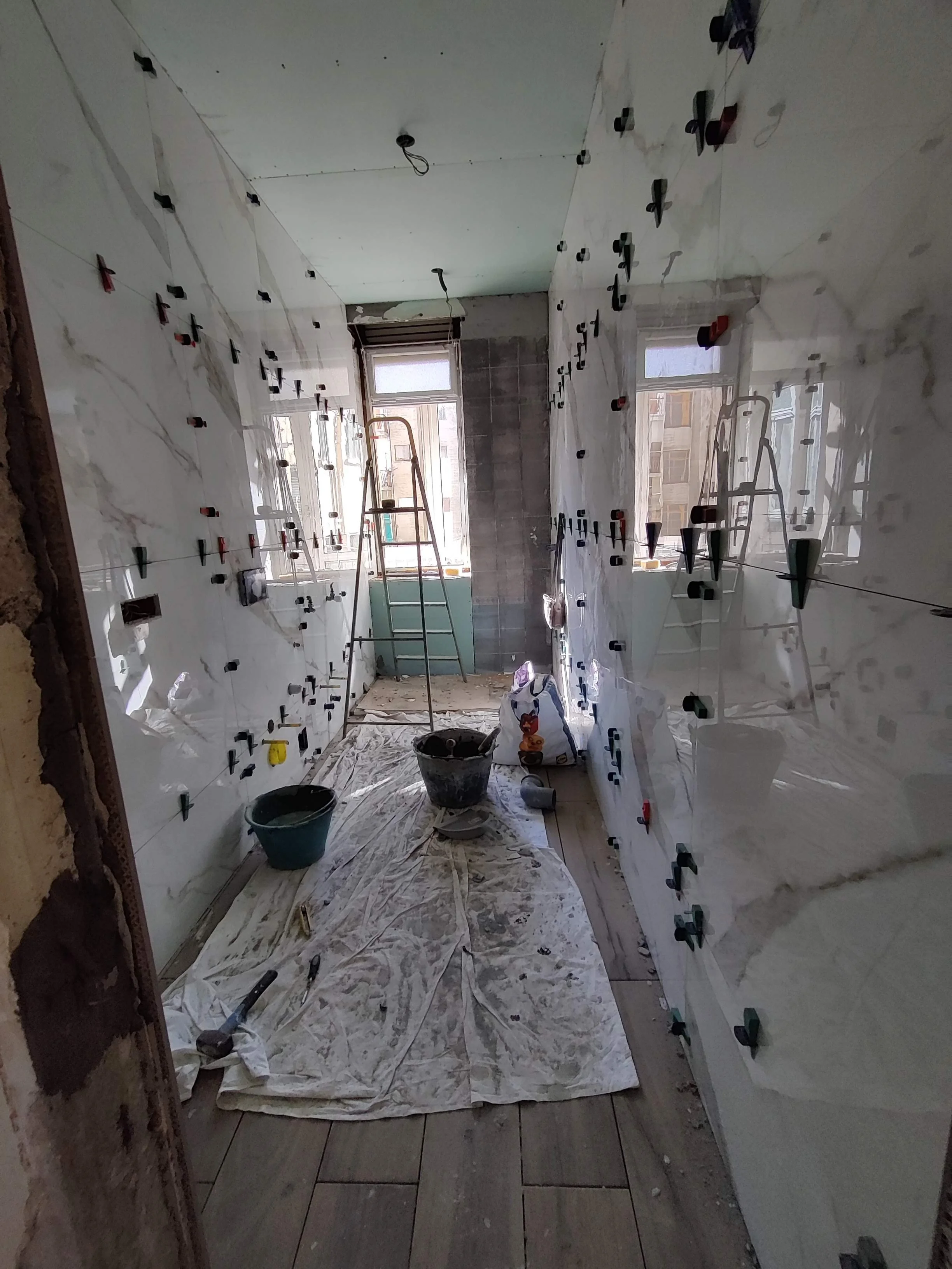 Interno di una stanza in fase di ristrutturazione con pareti rivestite di piastrelle, alcune parti ancora in lavorazione, attrezzi e materiale da costruzione, scala, e due finestre che consentono l'ingresso di luce naturale.