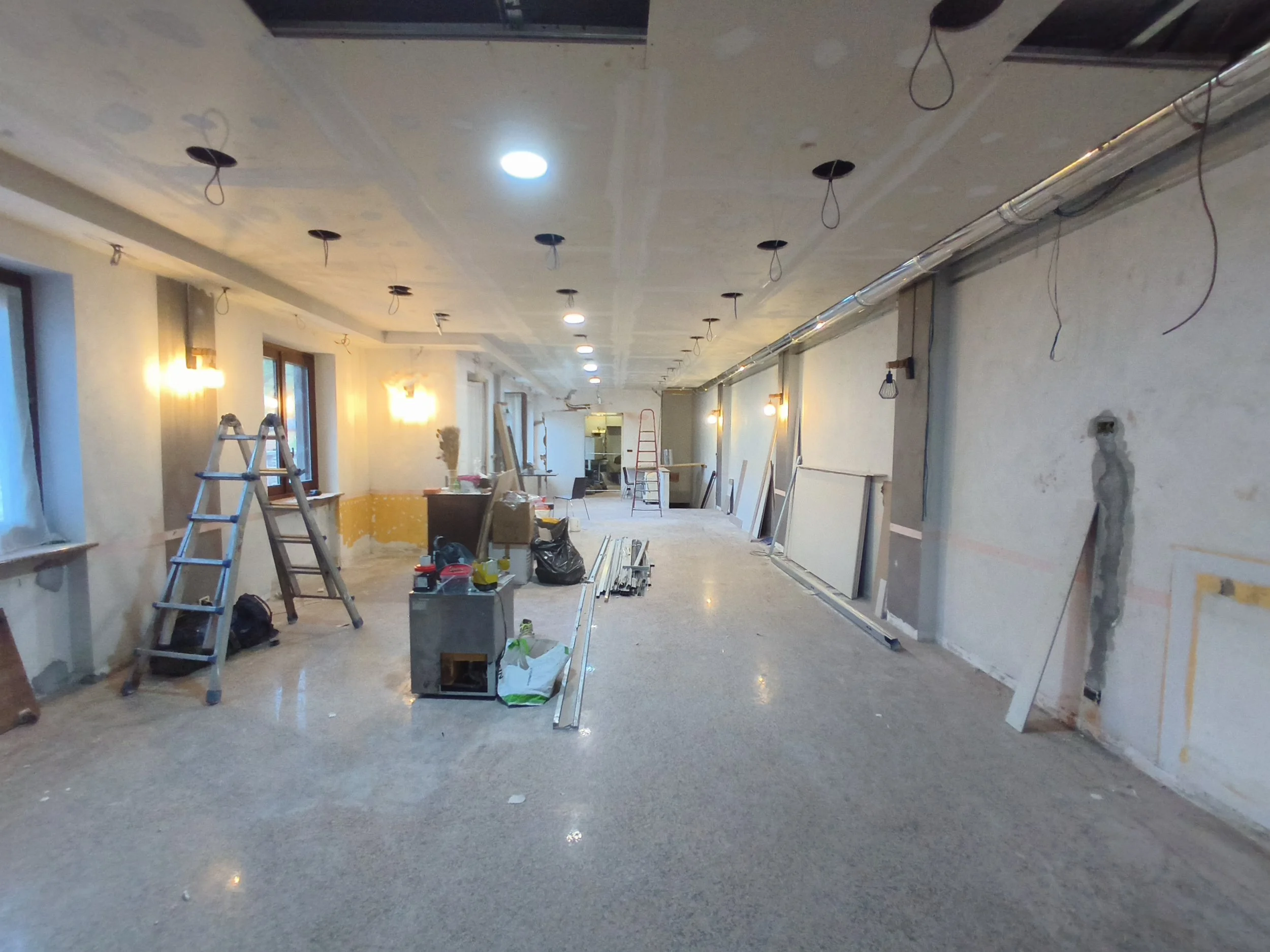 Interno di un edificio in fase di ristrutturazione con ponteggi, scala e materiali da costruzione, luci accese e pareti danneggiate.