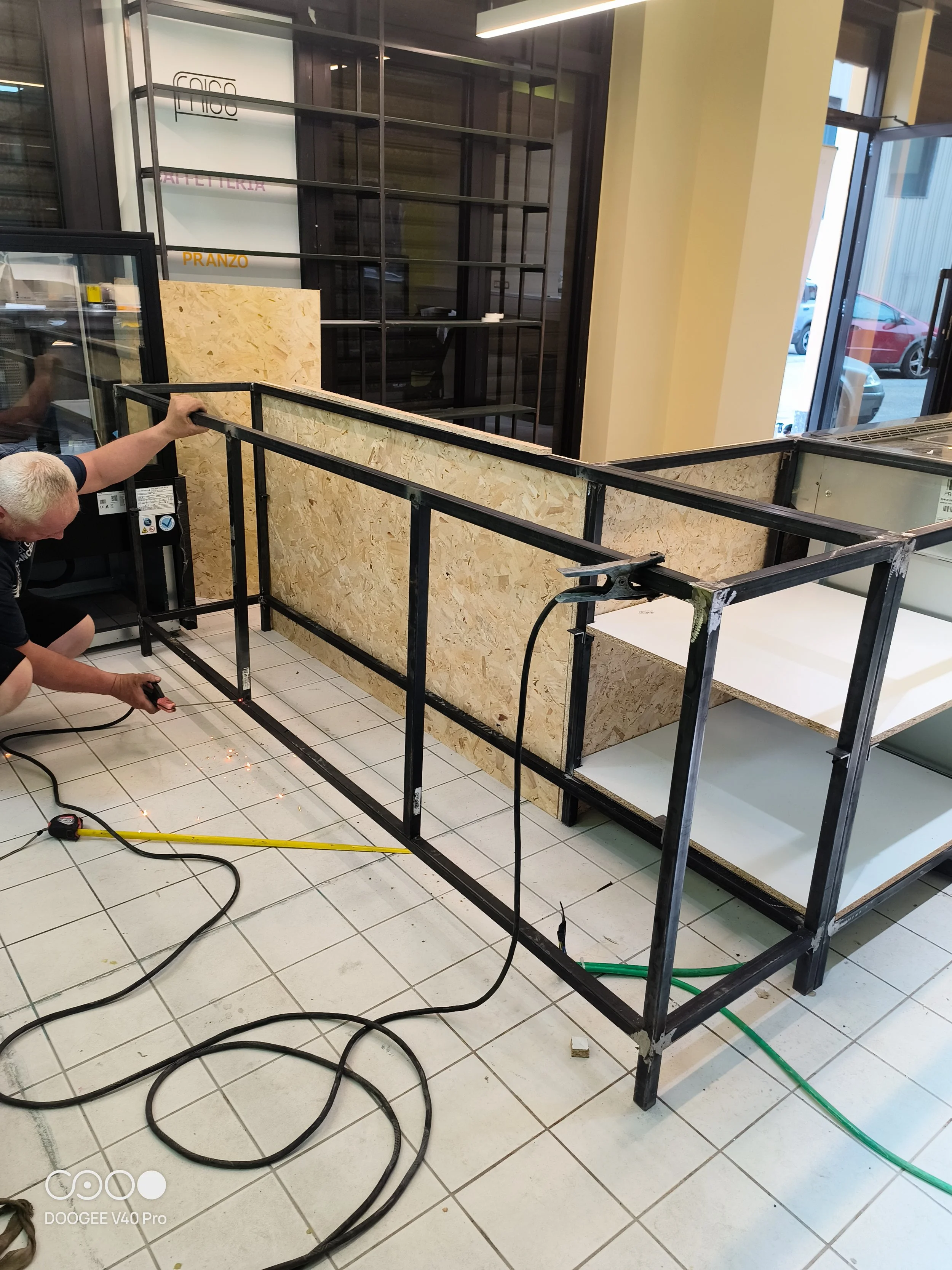 Personne travaillant à l'installation d'une structure métallique dans un commerce, avec des matériaux en bois et des câbles électriques sur le sol.