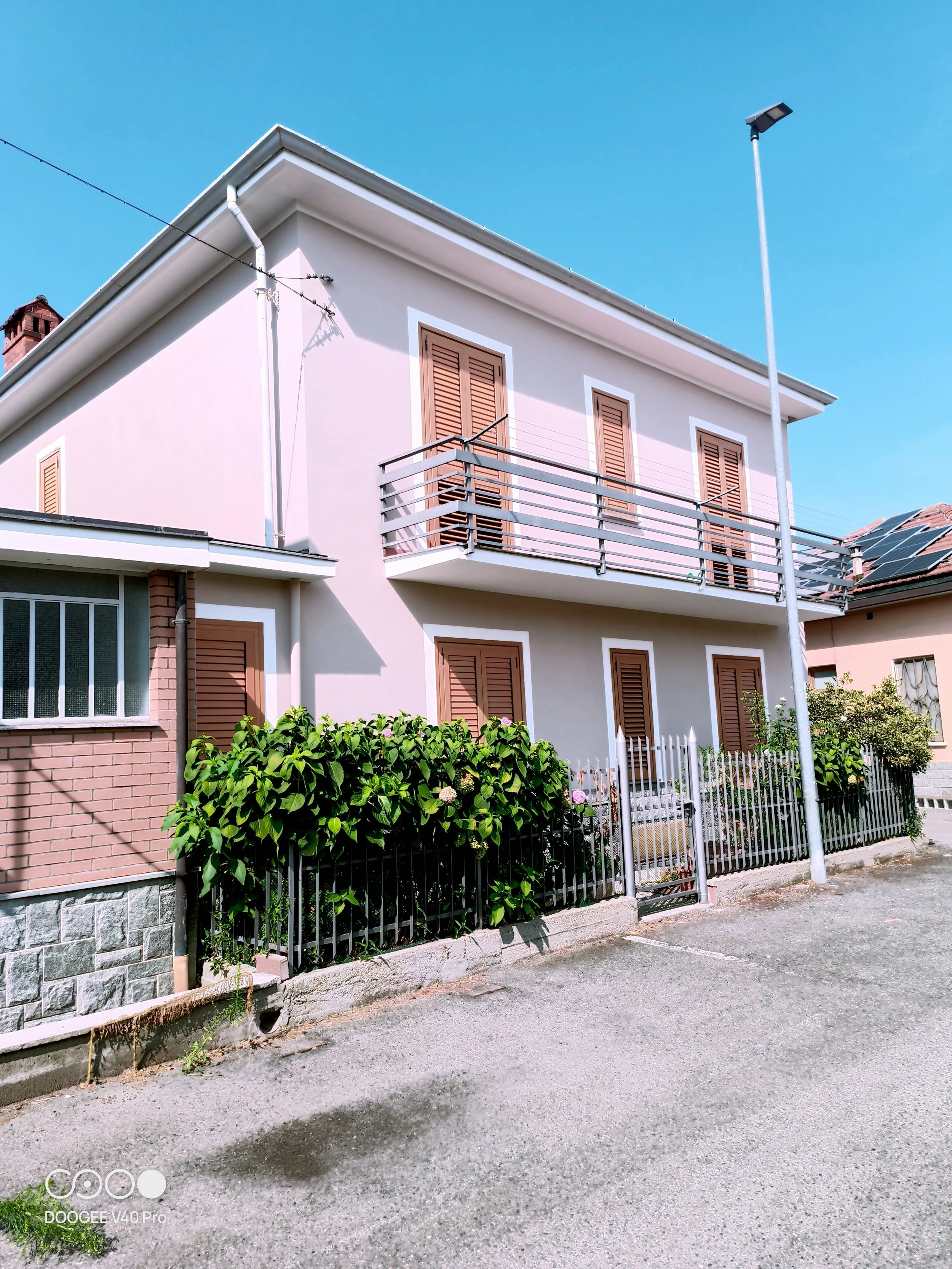 Edificio residenziale con pareti bianche, finestre con tapparelle di legno e un balcone con ringhiera di metallo. In giardino ci sono arbusti verdi e fiori, e una recinzione di metallo che delimita il cortile. Il cielo è azzurro con alcune nuvole sparse.