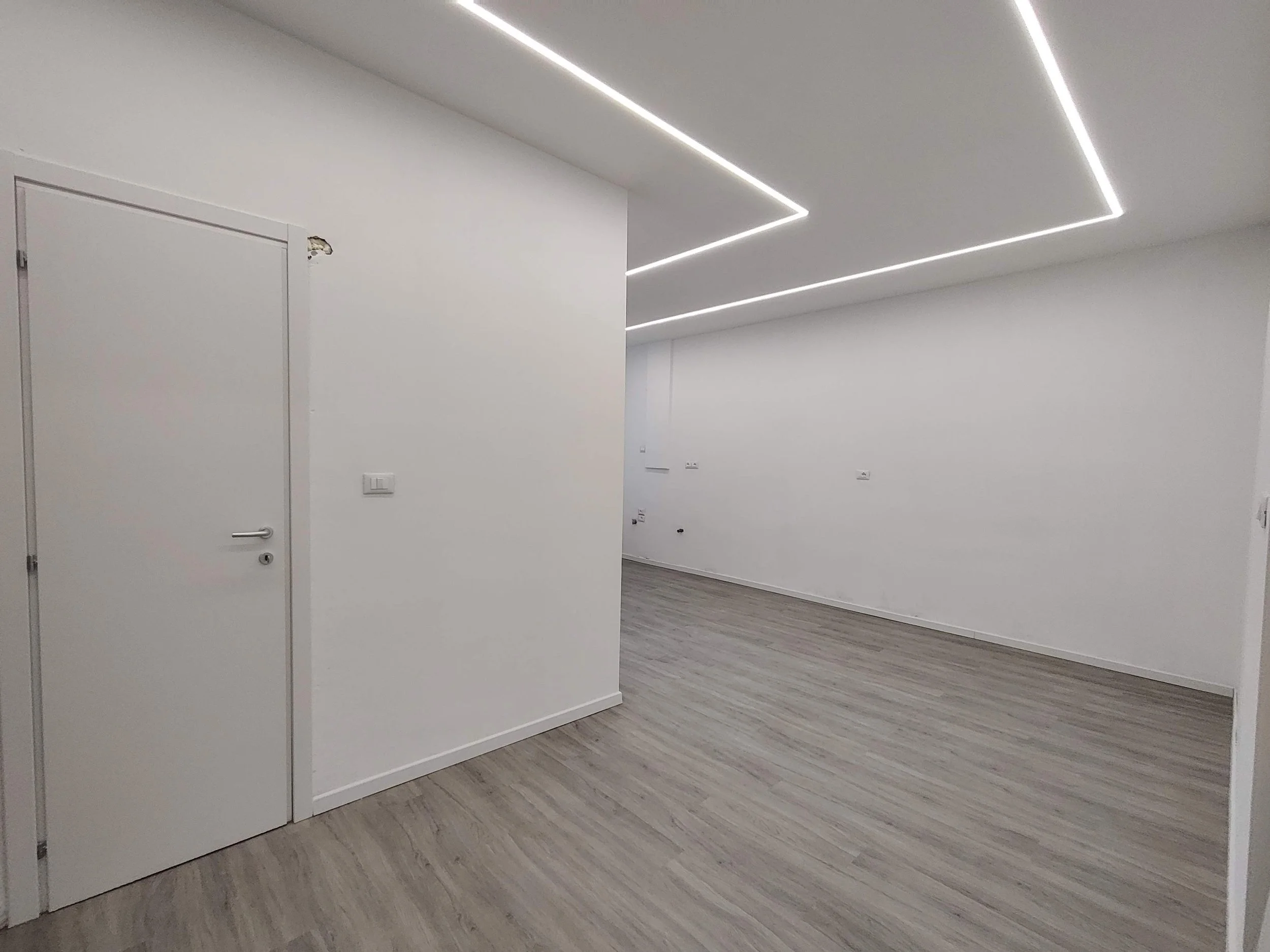 Interno di una stanza moderna con pareti bianche, pavimento in legno chiaro e un soffitto con illuminazione a LED disposta in linee parallele.
