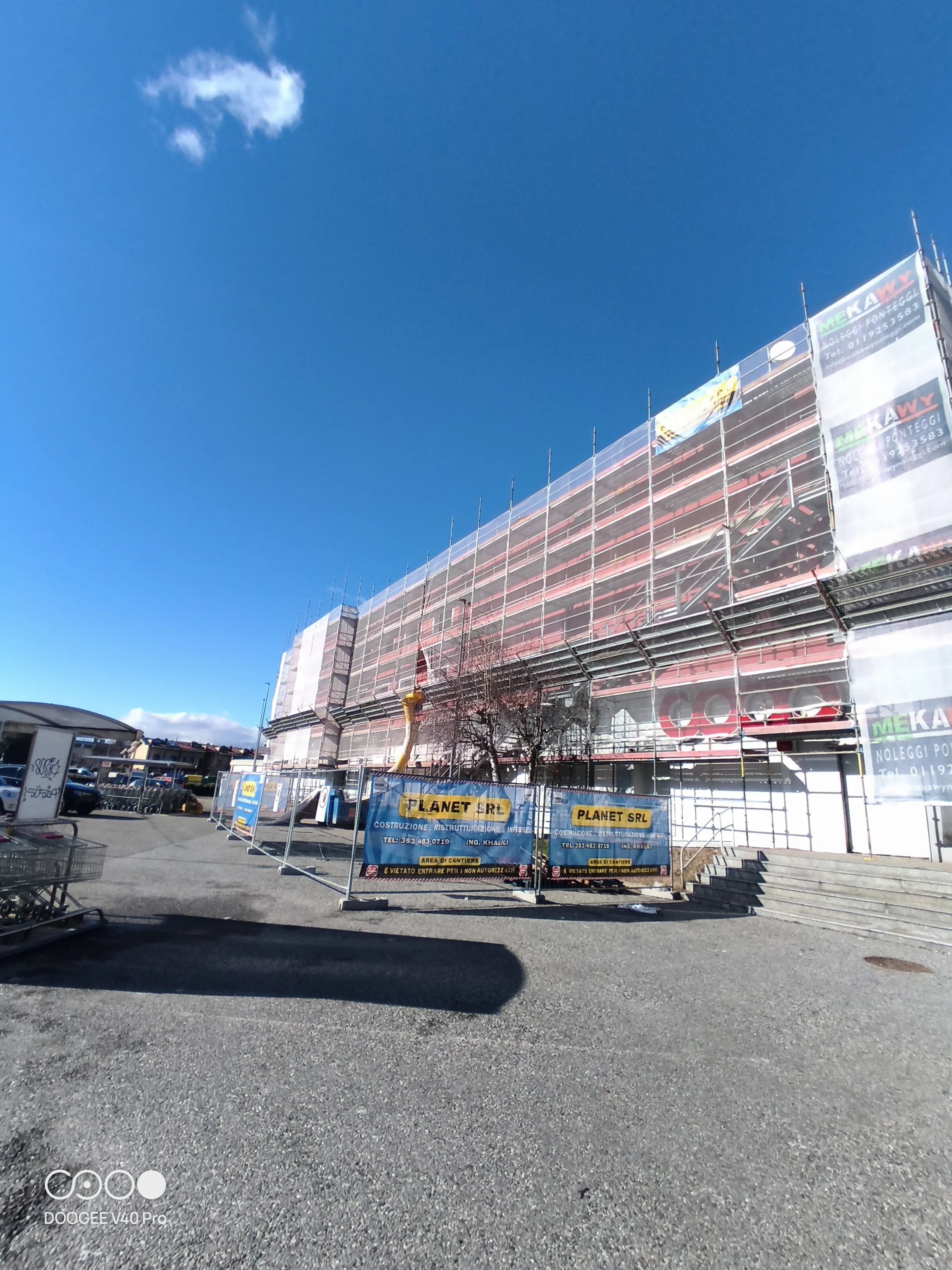 Cantiere di costruzione con recinzioni e segnali di sicurezza davanti a un edificio in ristrutturazione, sotto un cielo blu con poche nuvole.