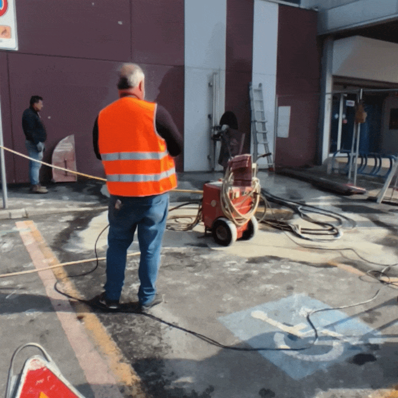 Operai lavorano alla riparazione di una superficie asfaltata con attrezzature e cavi, vicino a un edificio con dipinte di viola e una rampa di accesso.