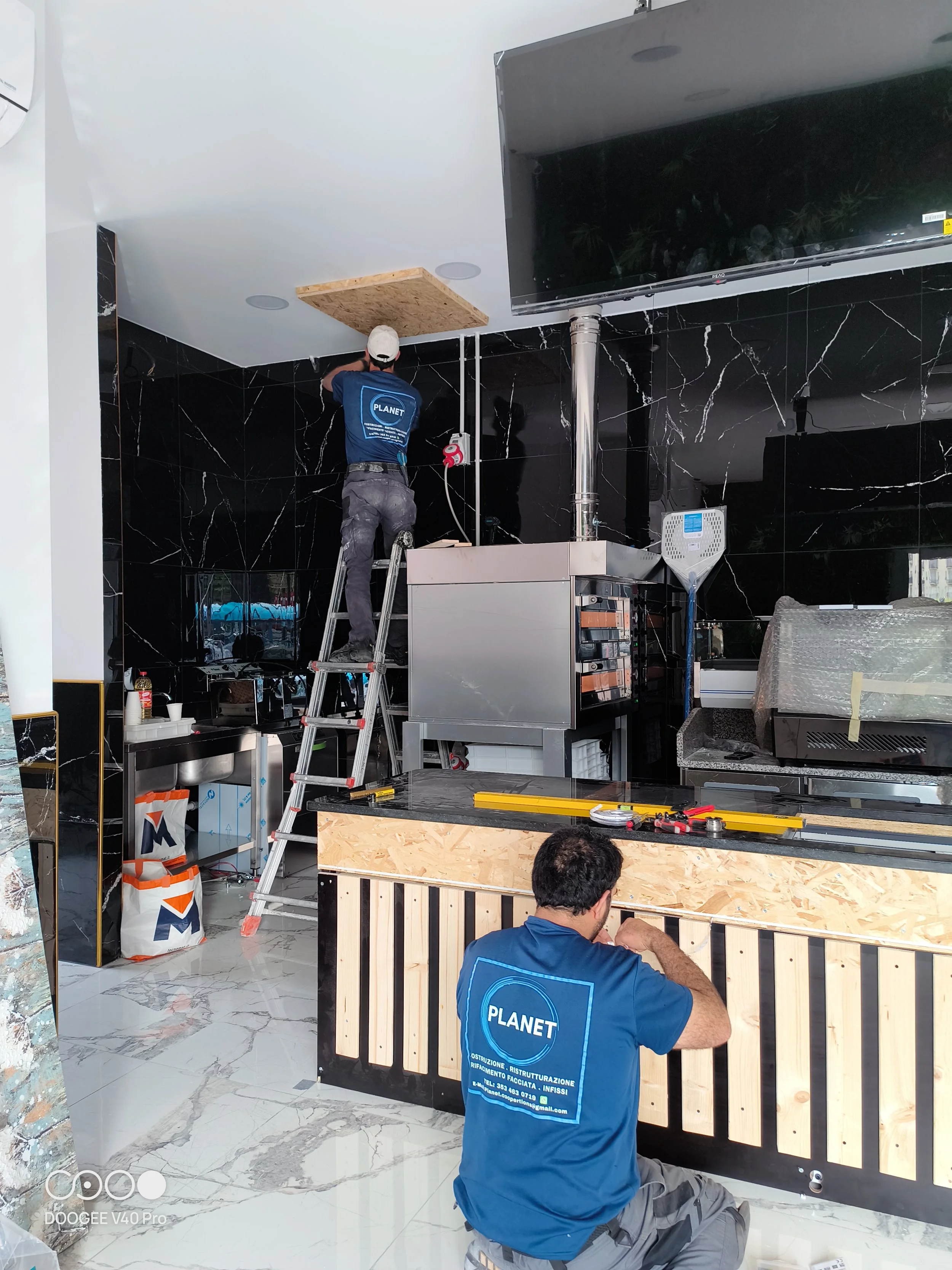 Due lavoratori installano un soffitto e una parete in un ambiente commerciale o ristorante. Uno di loro sta salendo su una scala, mentre l'altro lavora sulla parete in legno.