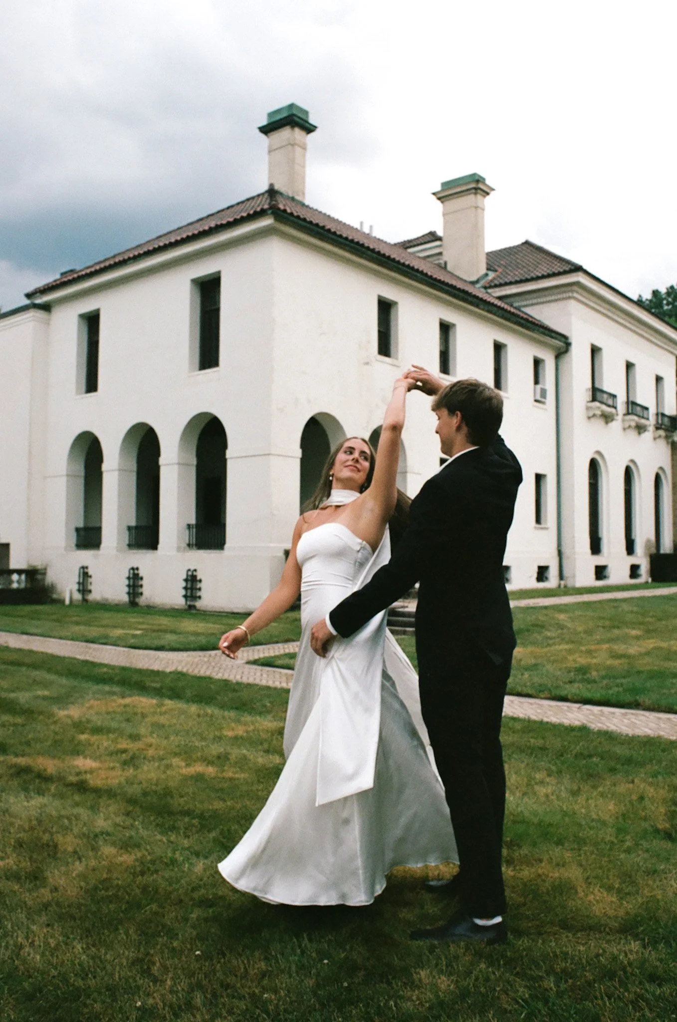 A couple dancing outdoors on grass in front of a large white building with arched windows and chimney stacks, dressed in formal wedding attire.