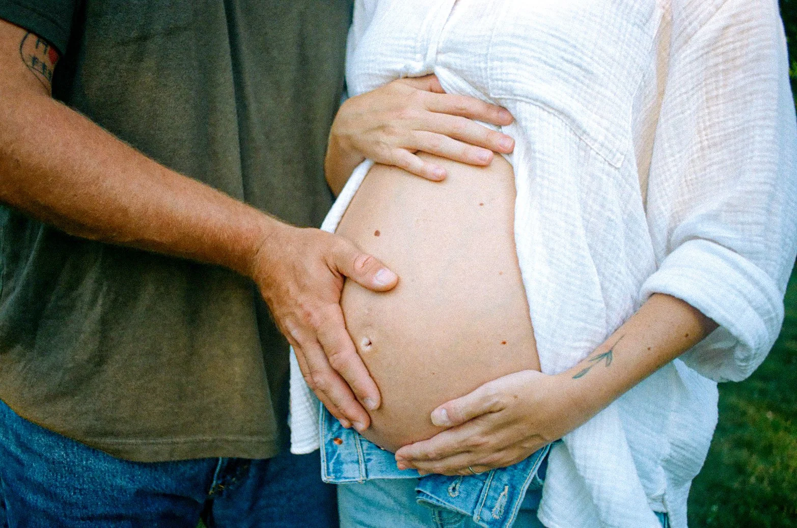 A pregnant woman holding her belly with her hands, standing outdoors, with a man partially visible next to her, touching her belly in a gentle manner.