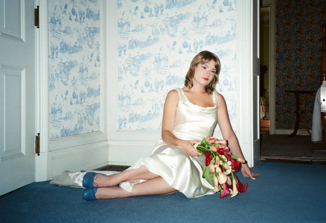 A woman in a white satin dress, with blue shoes, sitting on a blue carpeted floor, holding a bouquet of various colored flowers, in a room with white and blue patterned wallpaper and an open door leading to another room.