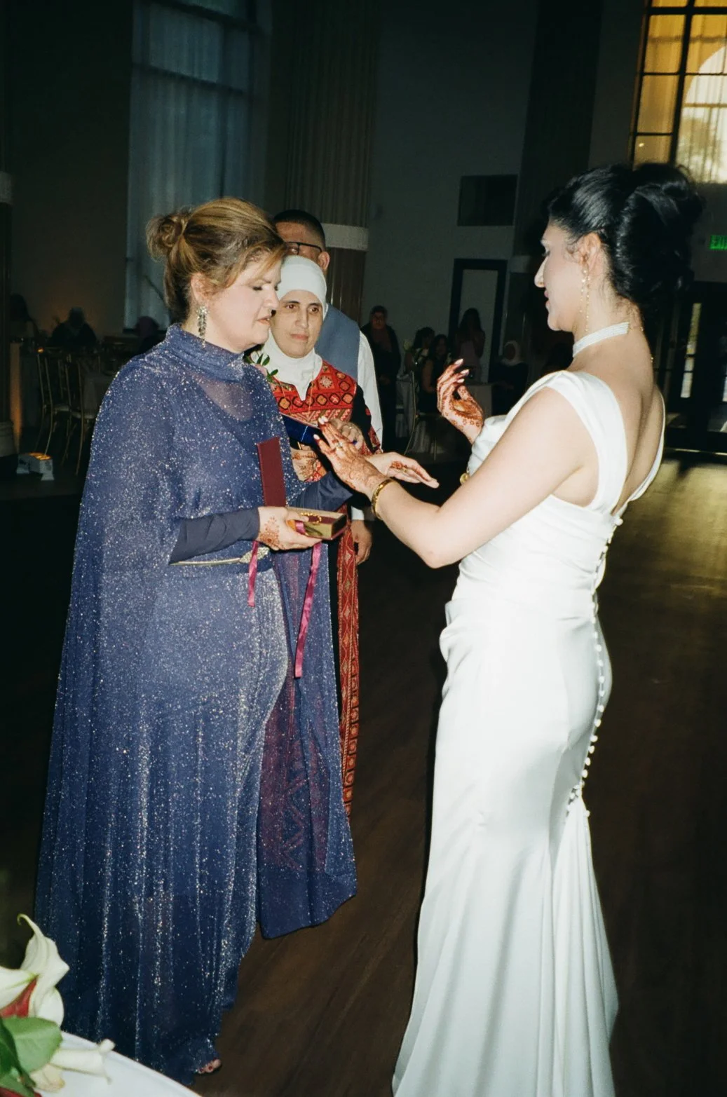 A bride in a white gown with buttons down the back and henna on her hands is presenting a gift to a woman in a glittery blue dress during a wedding ceremony, with a woman in traditional attire and a man in the background.