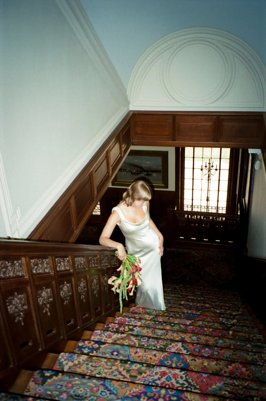 A woman in a white satin dress at the top of a wooden staircase, holding a bouquet of pink and white flowers, in a room with wooden paneling and a large window with a chandelier outside.
