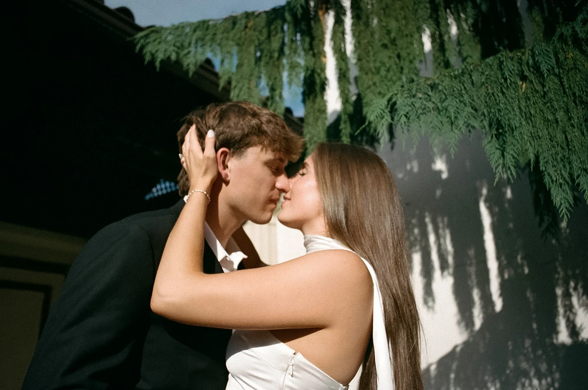 A young man and woman are kissing outdoors next to a large green tree, with their eyes closed and arms around each other.