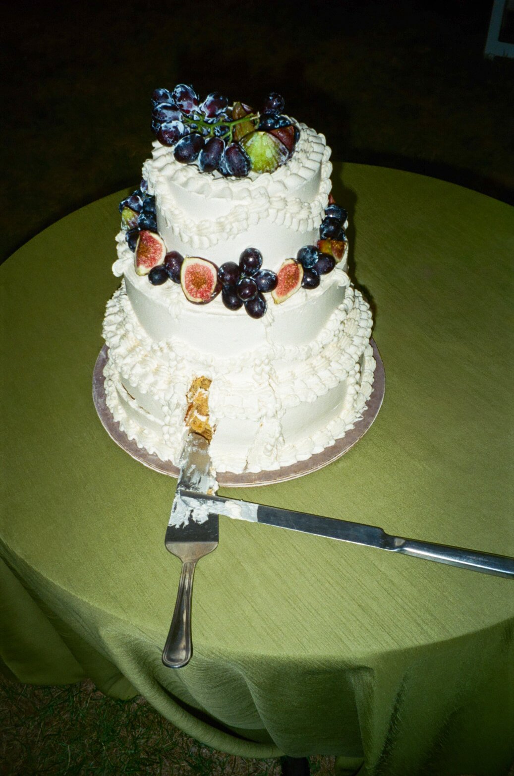 A three-tiered white wedding cake decorated with fresh grapes, figs, and whipped cream, with a slice already cut and a serving spatula on a green tablecloth.