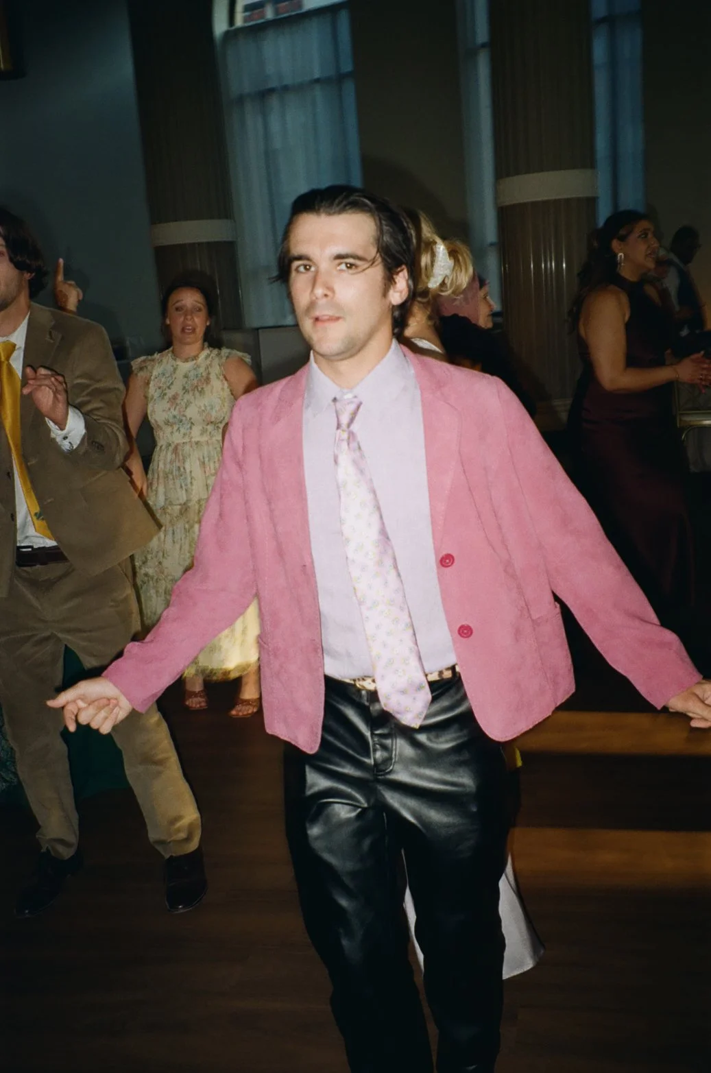 A man with dark hair, wearing a pink blazer, light-colored patterned tie, light shirt, and leather pants, dancing at a social event with other people in the background.