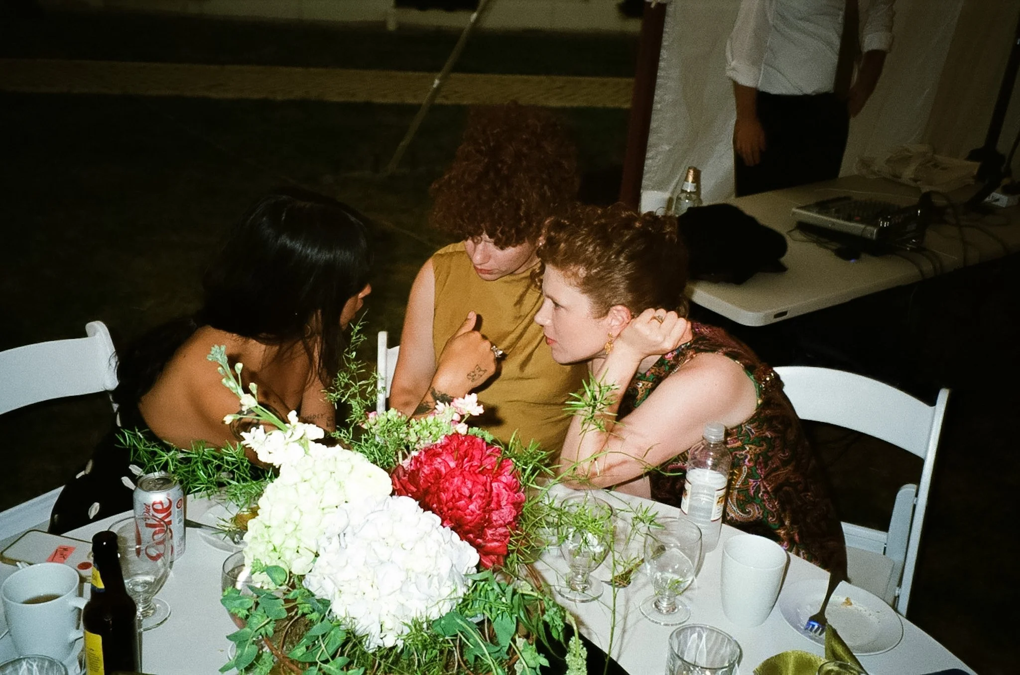 Three women with light to medium skin tones seated at a table, engaged in conversation, with a large floral centerpiece of red, white, and green flowers, surrounded by various drinks and bottles. They are indoors, near a white wall, with a table visi