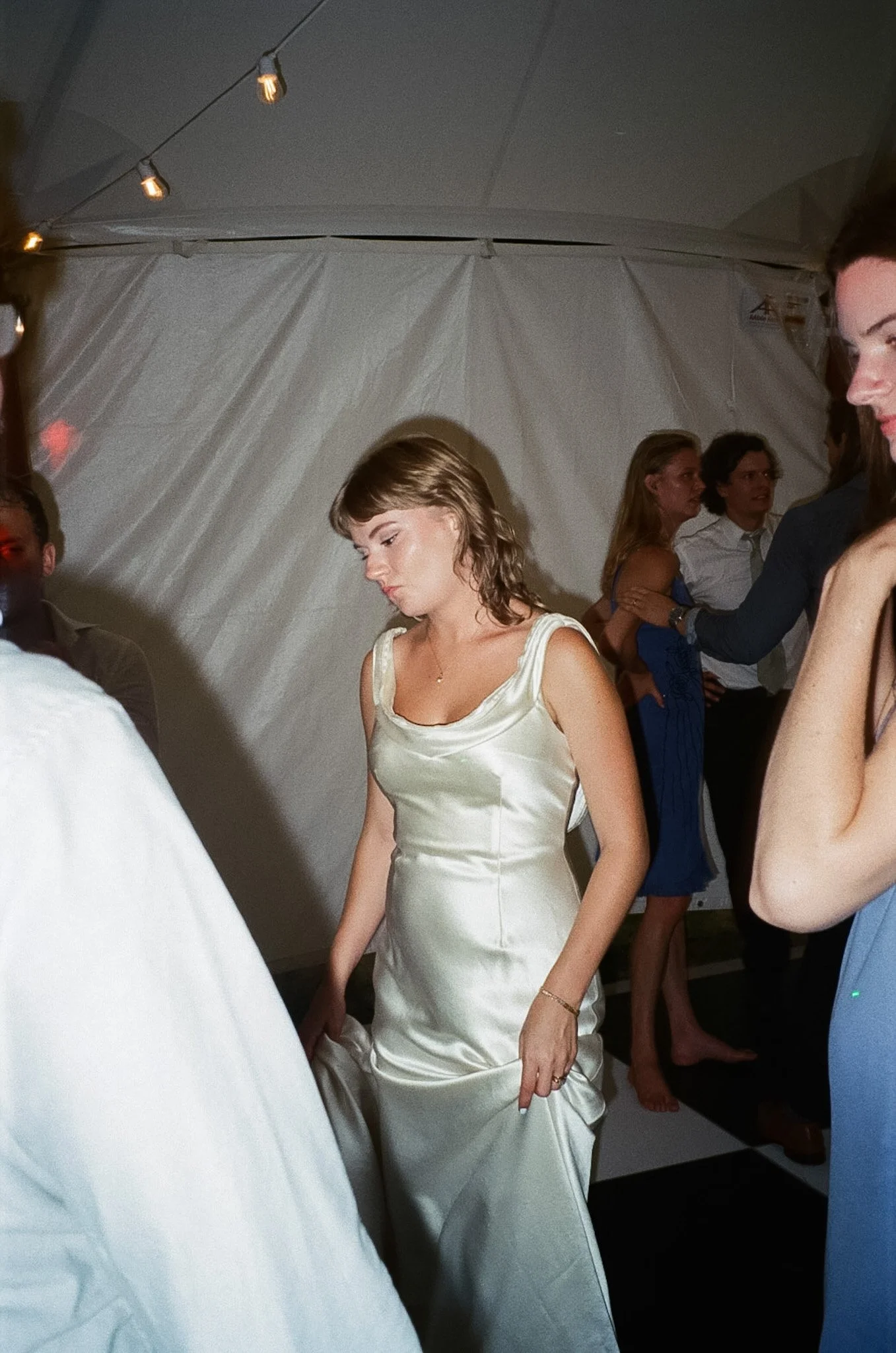 A woman in an elegant satin dress with thin straps, walking with her eyes down, in a room with white walls and string lights, surrounded by other well-dressed people at what appears to be a social gathering or party.