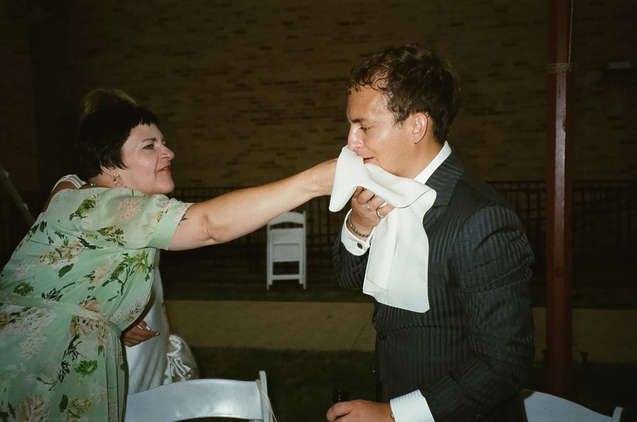 A woman is wiping a tear from a young man's face with a white cloth during a emotional moment at an outdoor event at night.