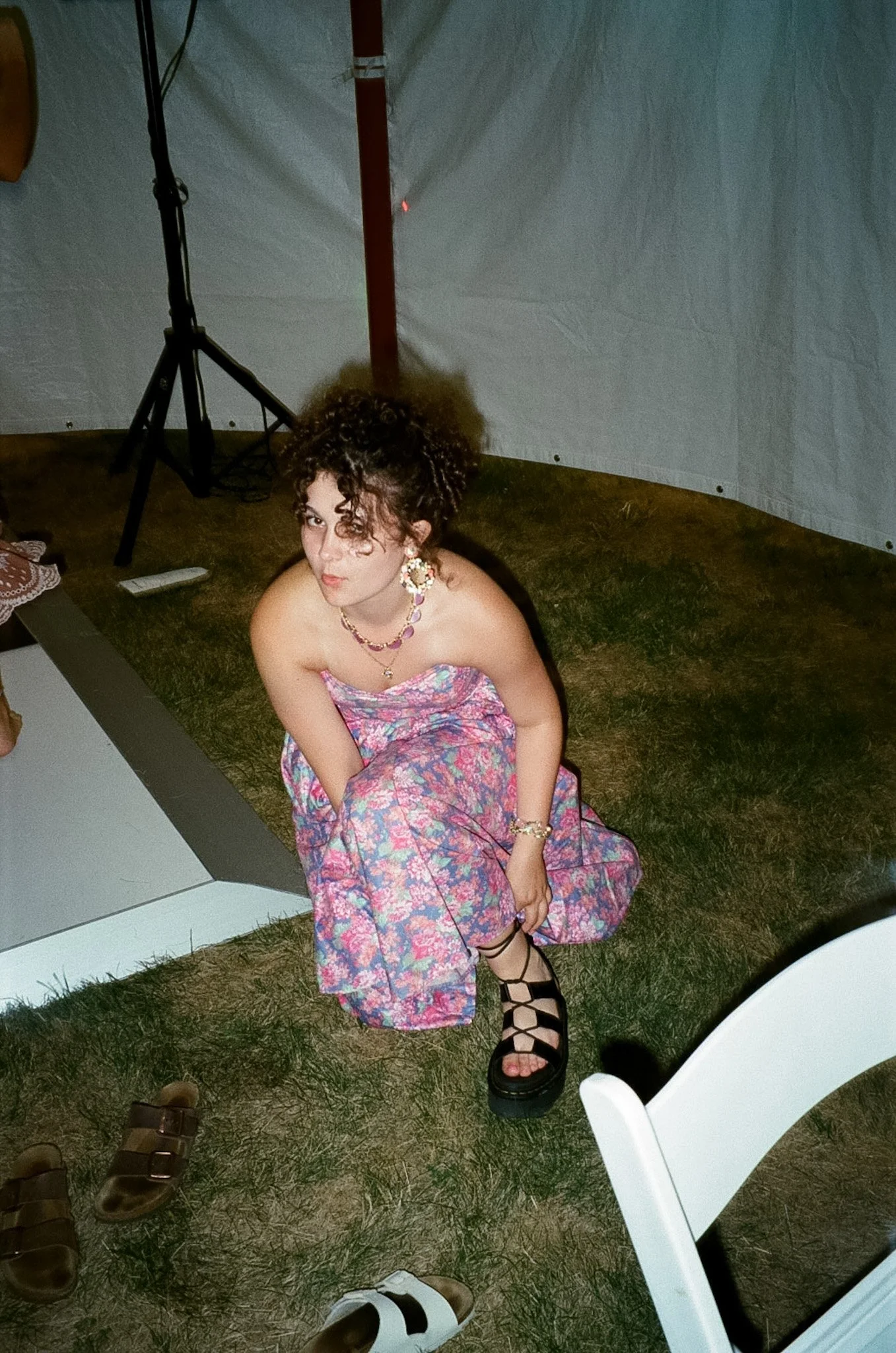 A woman with curly hair and hoop earrings wearing a colorful strapless dress with a floral pattern, sitting on the ground with her knees bent. She is wearing black lace-up sandals. Surrounding her are various pairs of shoes, including brown sandals, 